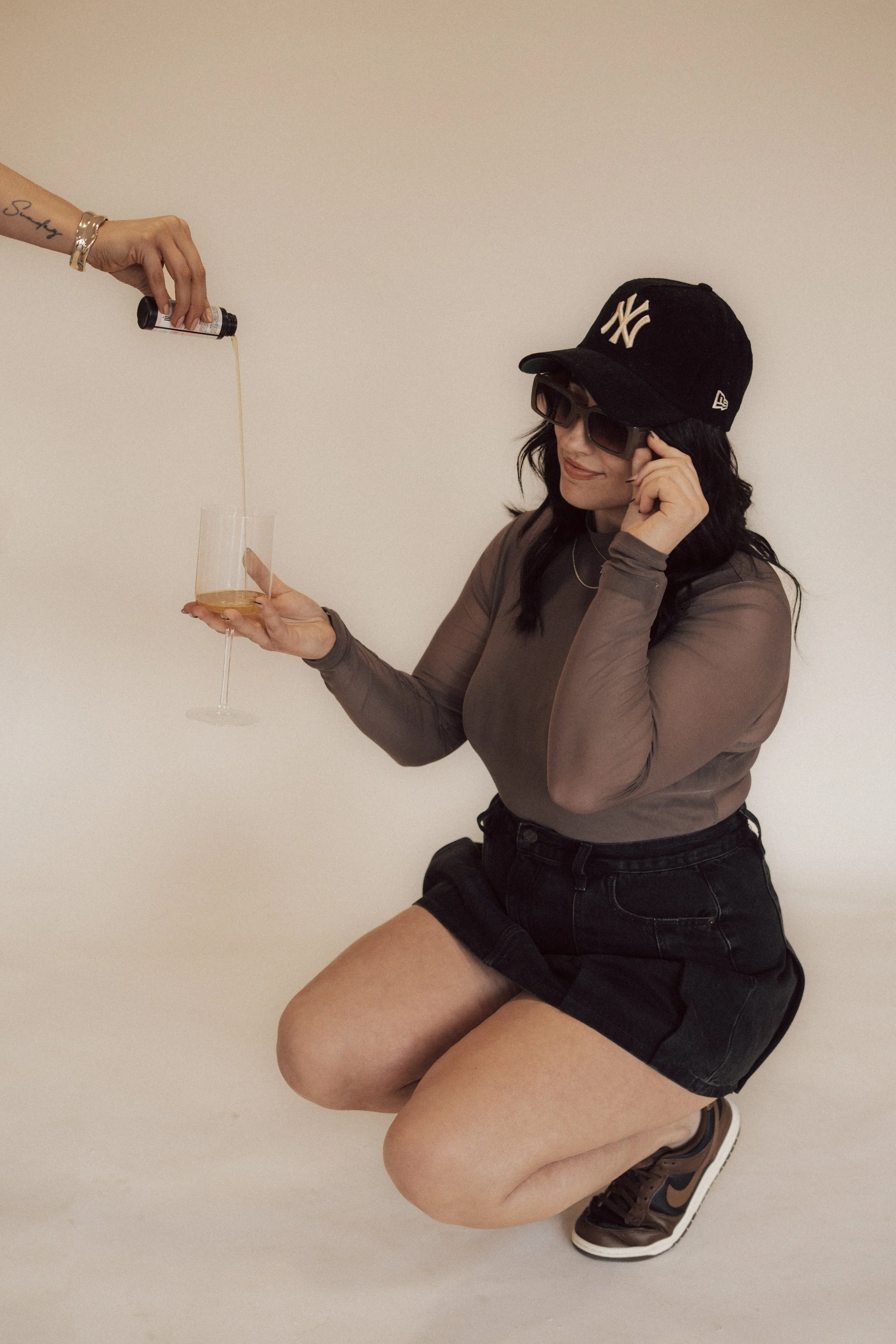 A woman in a black baseball cap, sunglasses, and a long-sleeve brown shirt is kneeling on the floor. Someone is pouring champagne into her glass, and she is holding the glass while smiling.