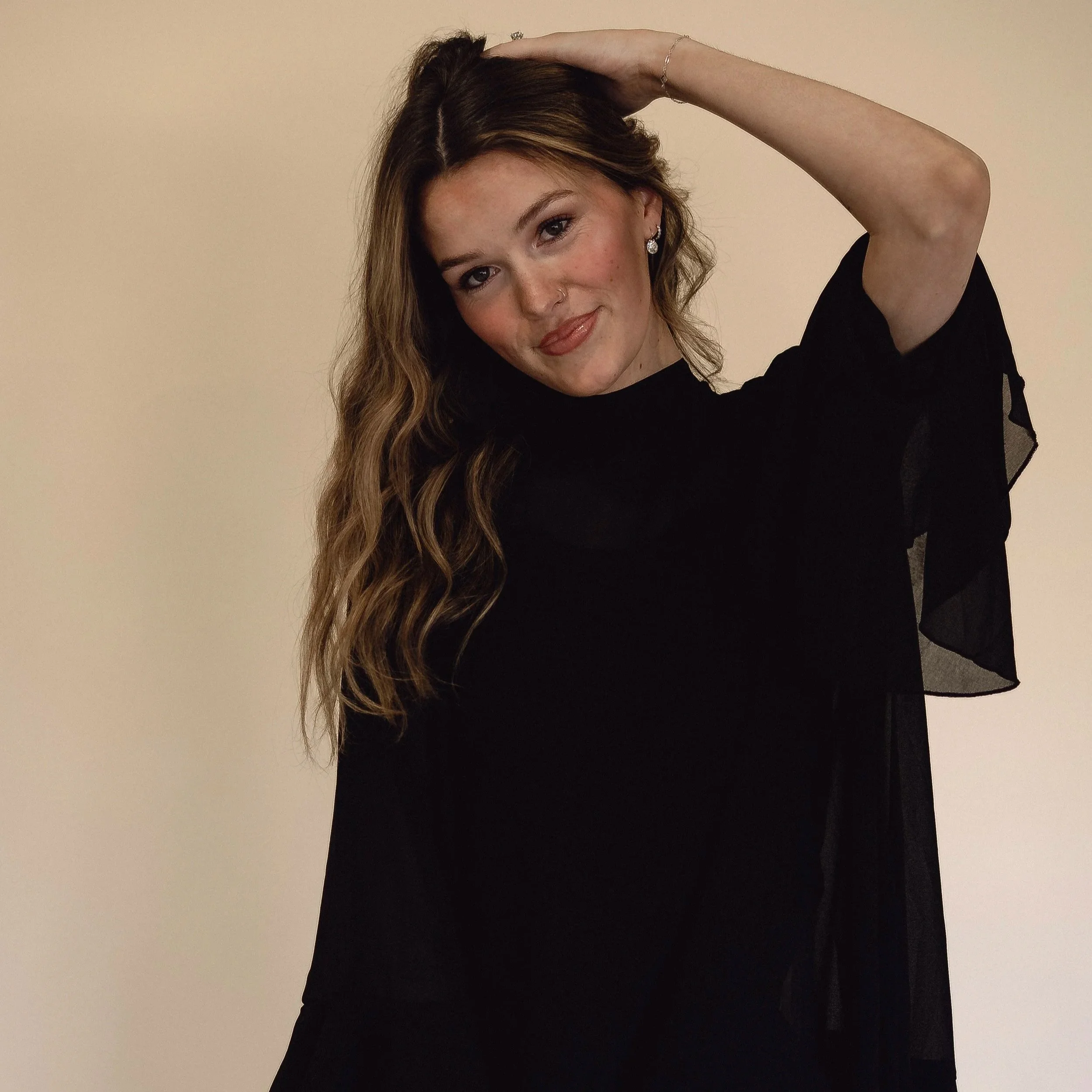 A woman with wavy brown hair and light skin, wearing a black dress with sheer sleeves, posing with her right hand resting on her head, smiling gently, against a plain light-colored background.