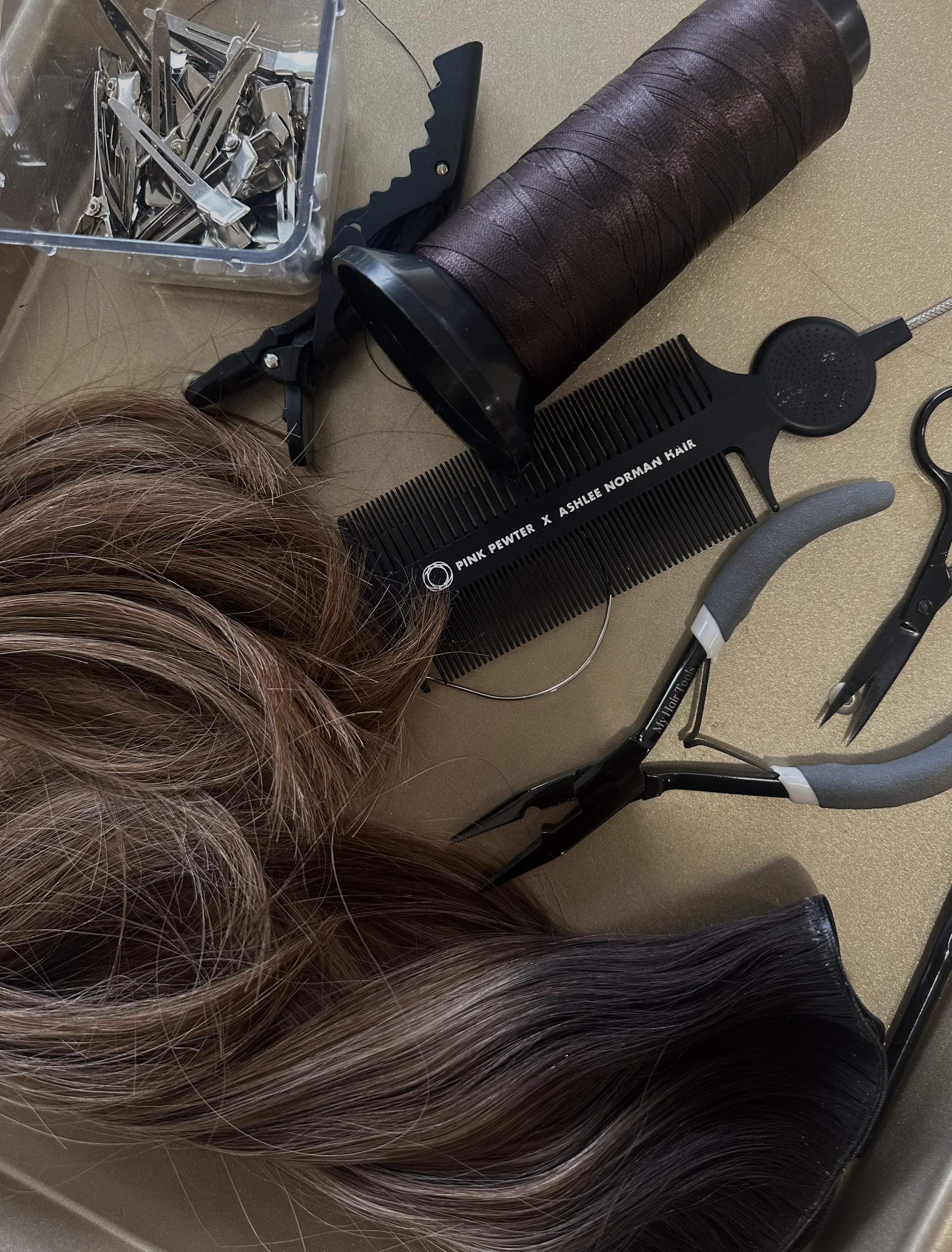 A collection of hairstyling tools including hair extensions, a large spool of brown hair, black metal clips, a wide black comb labeled "Pink Pewter x Ashlee Norman Hair," a pair of ratchet scissors, a rat tail comb with the label "My Hair Tools," a small plastic container with hair clips, and a pair of hair cutting shears on a beige surface.