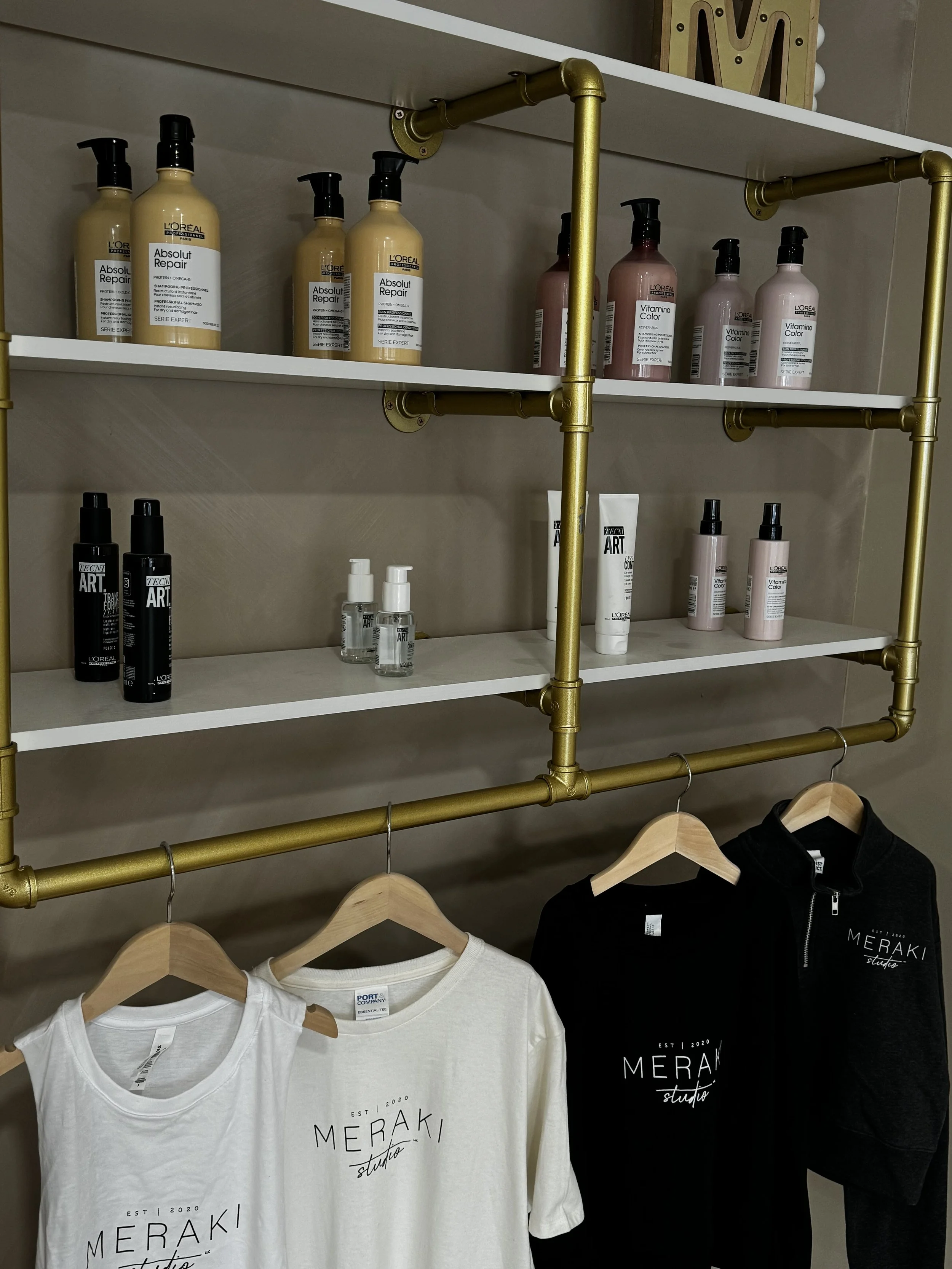 Display of hair care products on white shelves with black and pink bottles, and black spray bottles, above clothing items on wooden hangers including white and black T-shirts and a black zip-up hoodie, against a beige wall.