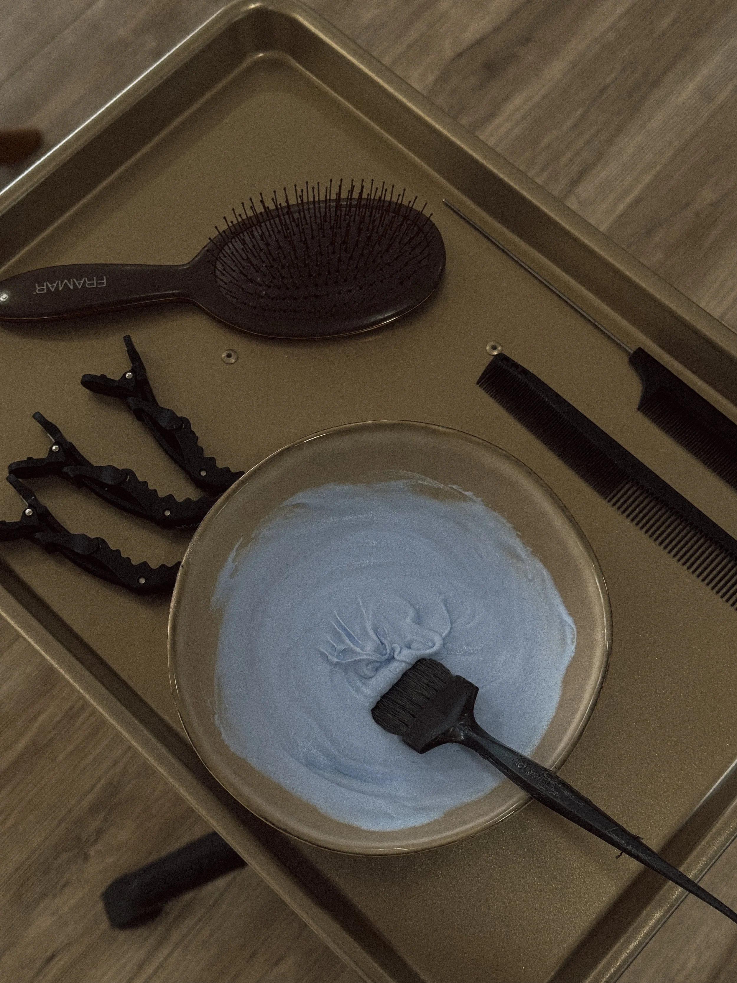 A bowl of light blue hair dye with a brush inside, a round hairbrush, three black hair clips, and two black combs on a gold-colored table.