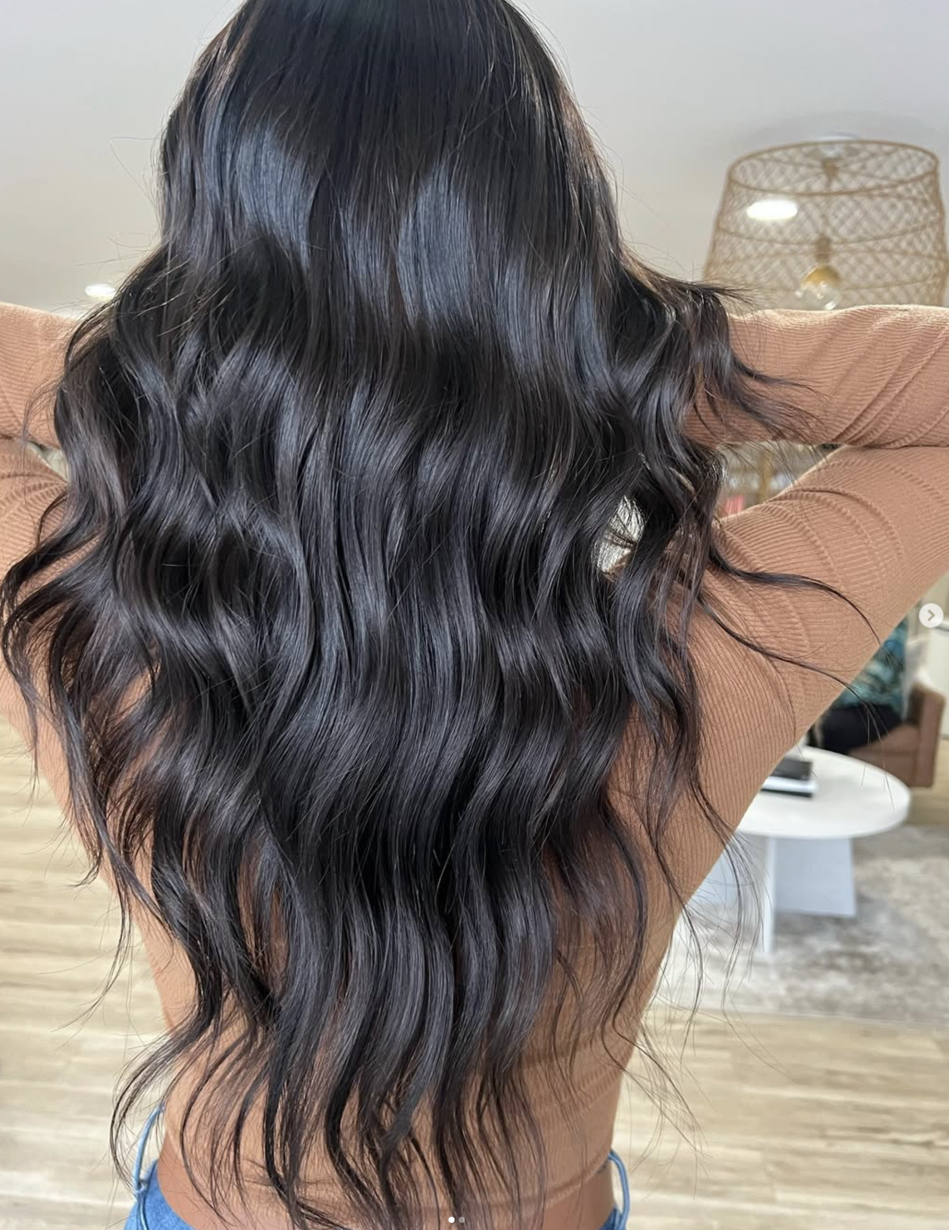 A person with long, wavy, dark brown or black hair, seen from behind, wearing a tan long-sleeve top and outdoors in a living room area.