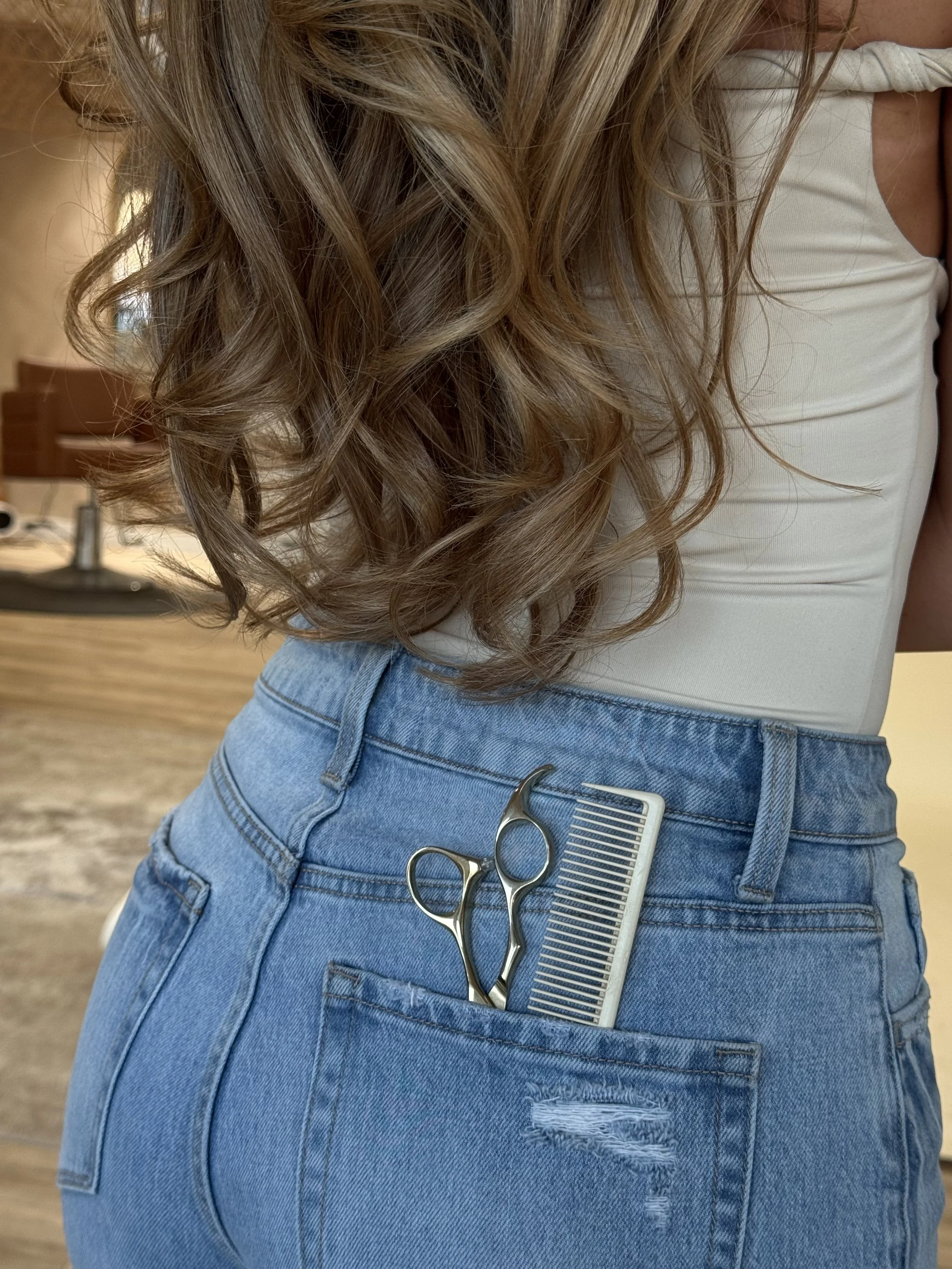 A person with long, wavy hair wearing a white tank top and high-waisted blue jeans, with hair cutting tools including scissors and a comb in the back pocket.