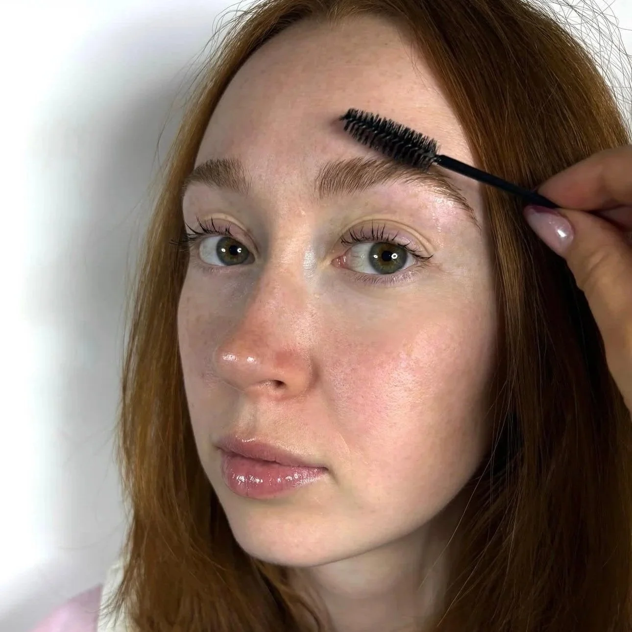 Woman with reddish hair getting her eyebrows brushed with a spoolie brush by another person.
