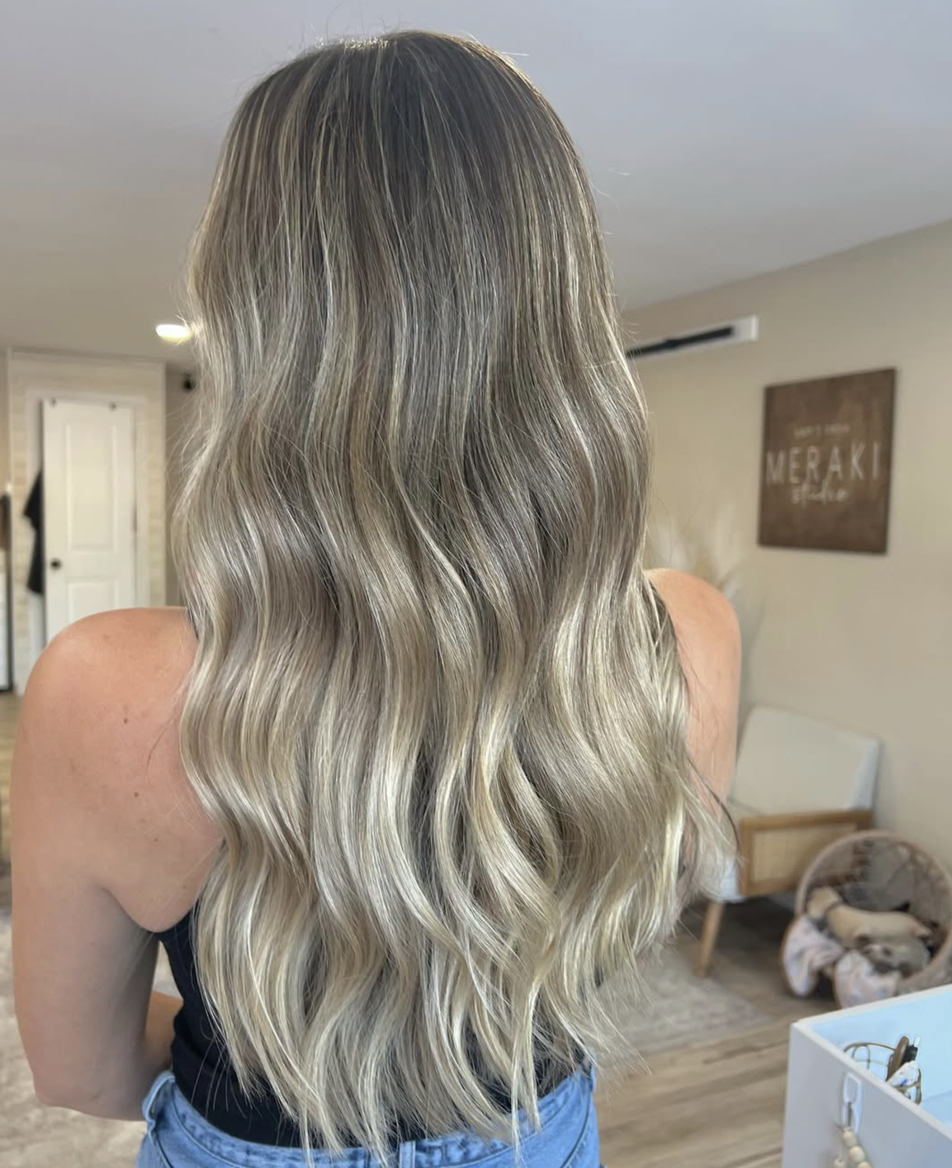 Back view of a woman with long, wavy blonde hair standing indoors.