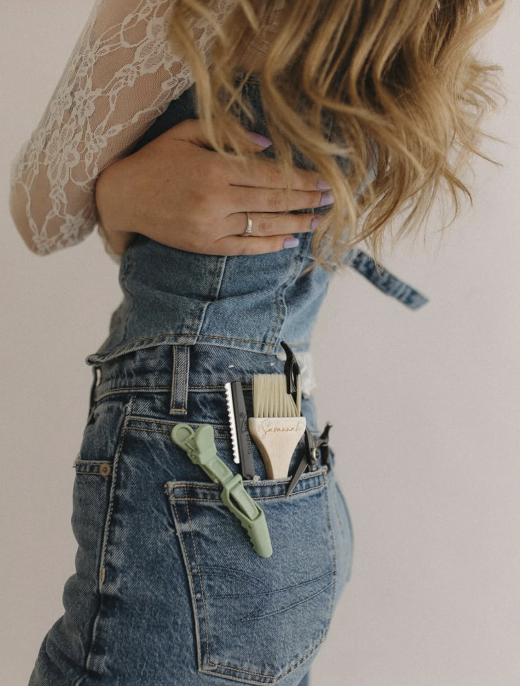 A woman with wavy blonde hair, wearing a lace long-sleeve top and high-waisted blue jeans, holds her side with one hand and has manicure nails.