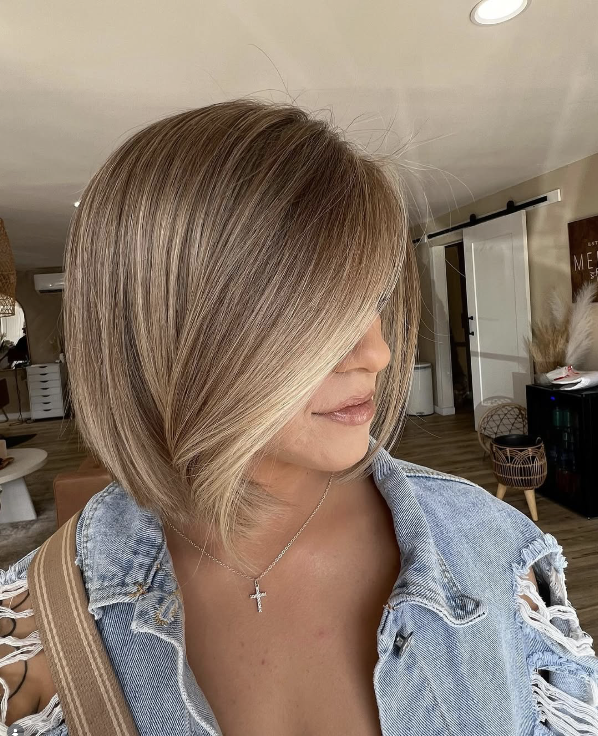 Woman with shoulder-length blonde layered hair, wearing denim jacket with distressed details and a cross necklace, in a cozy interior space.
