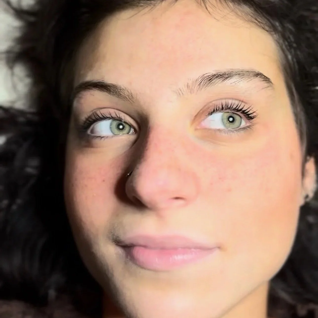 Close-up of a young woman's face with green eyes, dark hair, and smooth skin, looking slightly upward.