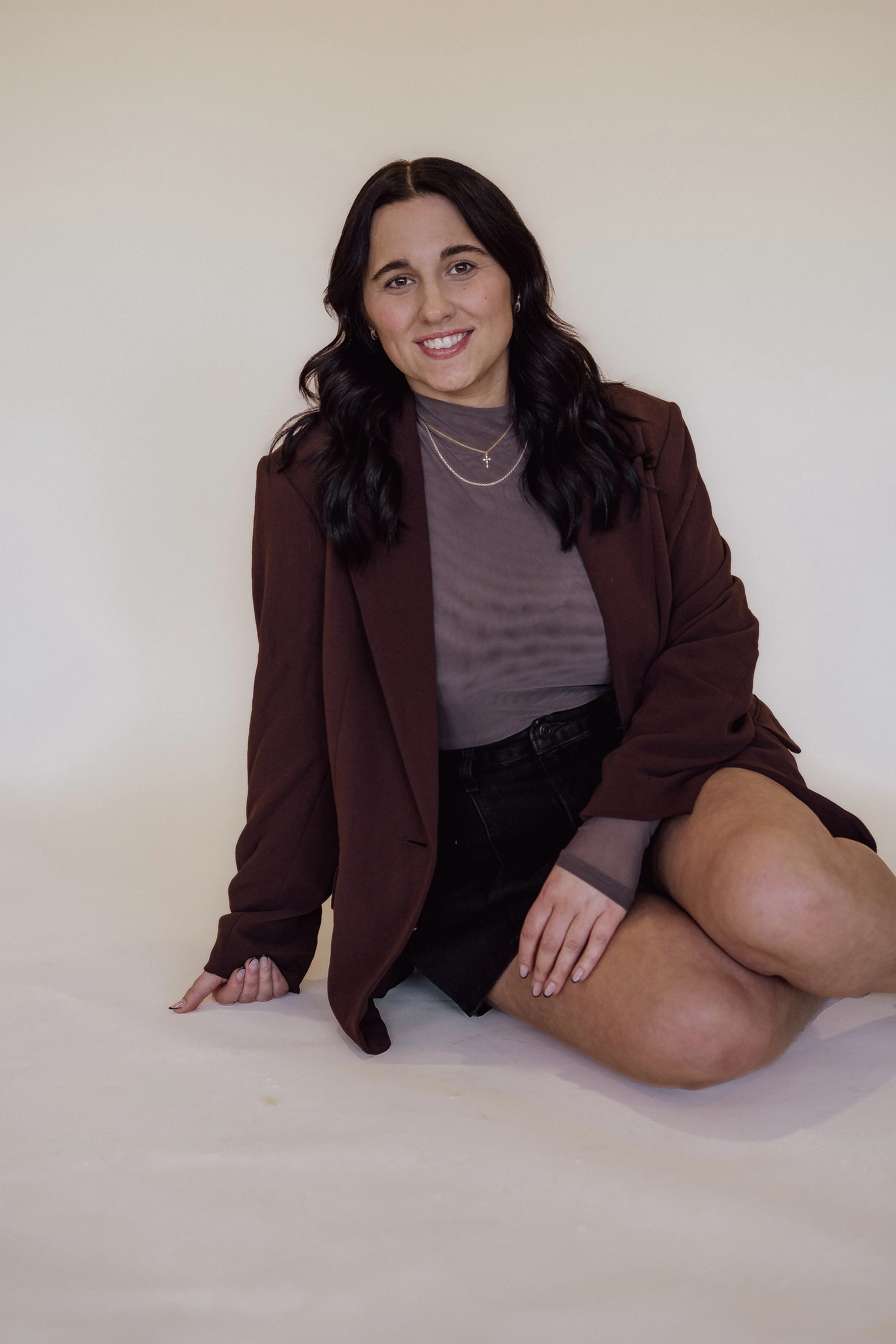 A woman with long dark hair, wearing a brown blazer, gray shirt, and black shorts, sitting on the floor against a plain light-colored wall, smiling at the camera.