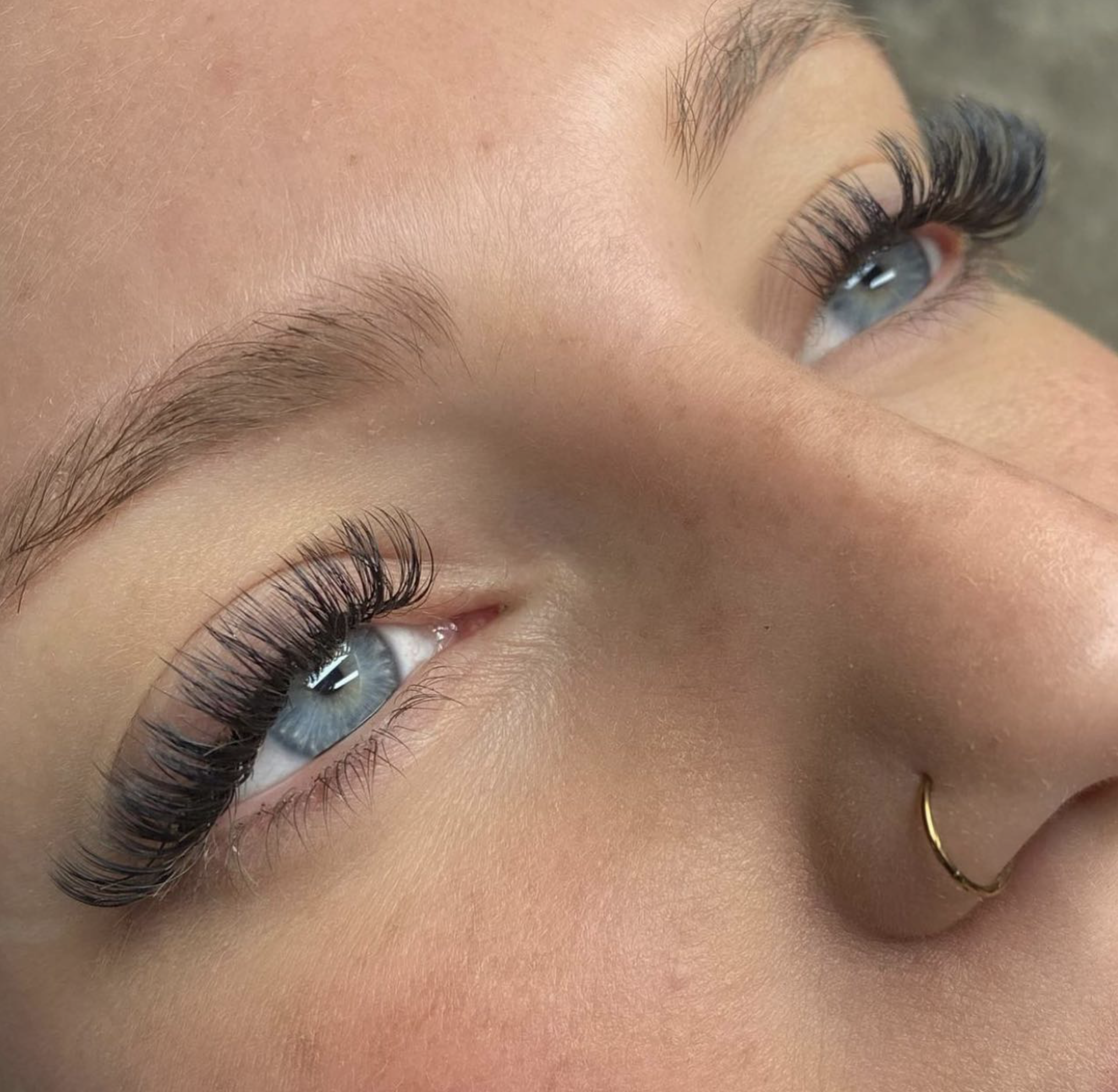 Close-up of a person's eyes showcasing long, curled eyelashes, blue irises, eyebrows, a nose piercing, and smooth skin.