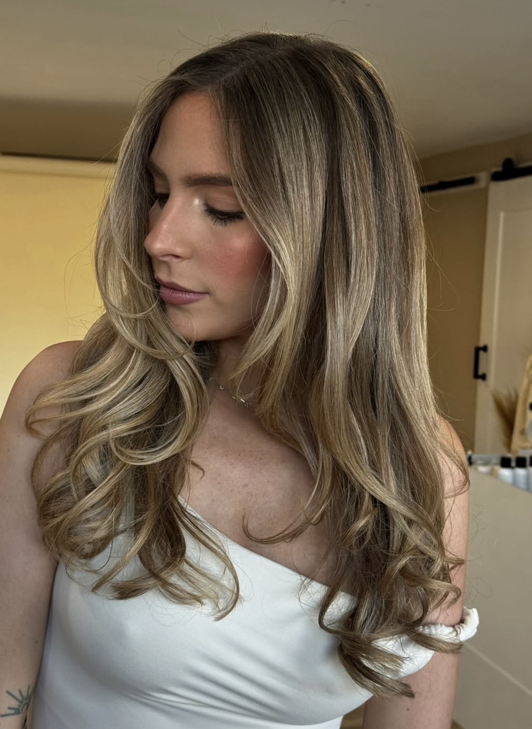 A young woman with long, wavy, blonde hair looking down, wearing a white sleeveless top, in a room with beige walls and a white door in the background.
