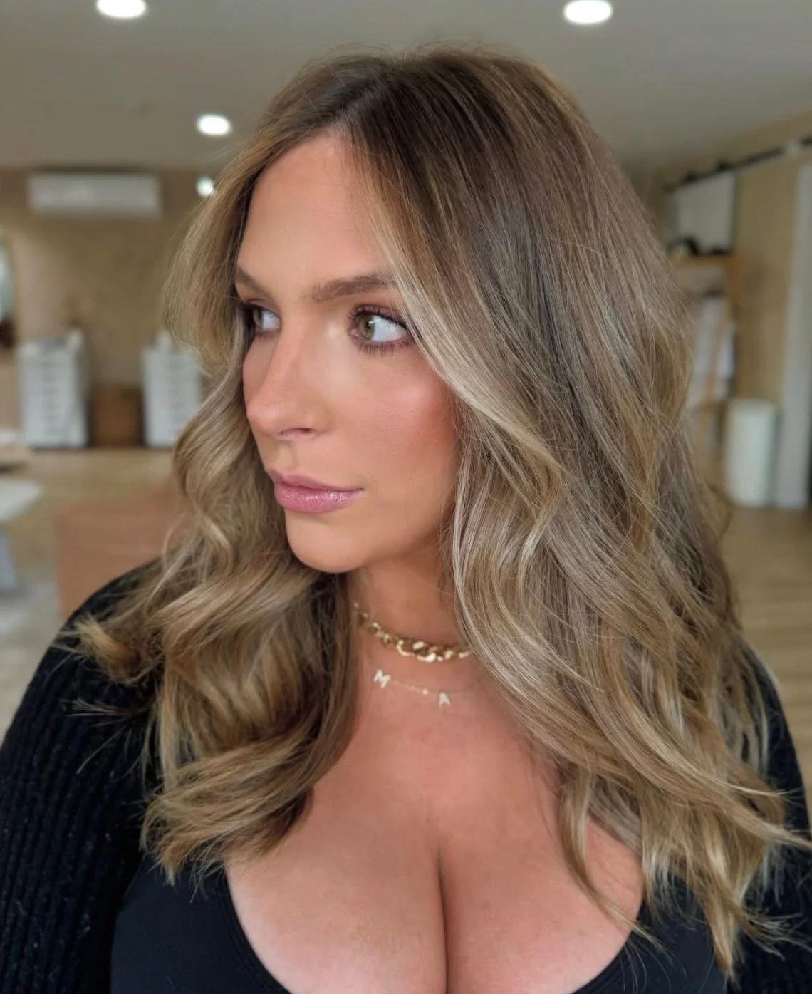 A woman with wavy, blonde hair and green eyes looking to her left, wearing layered necklaces and a black top, indoors with blurred background.