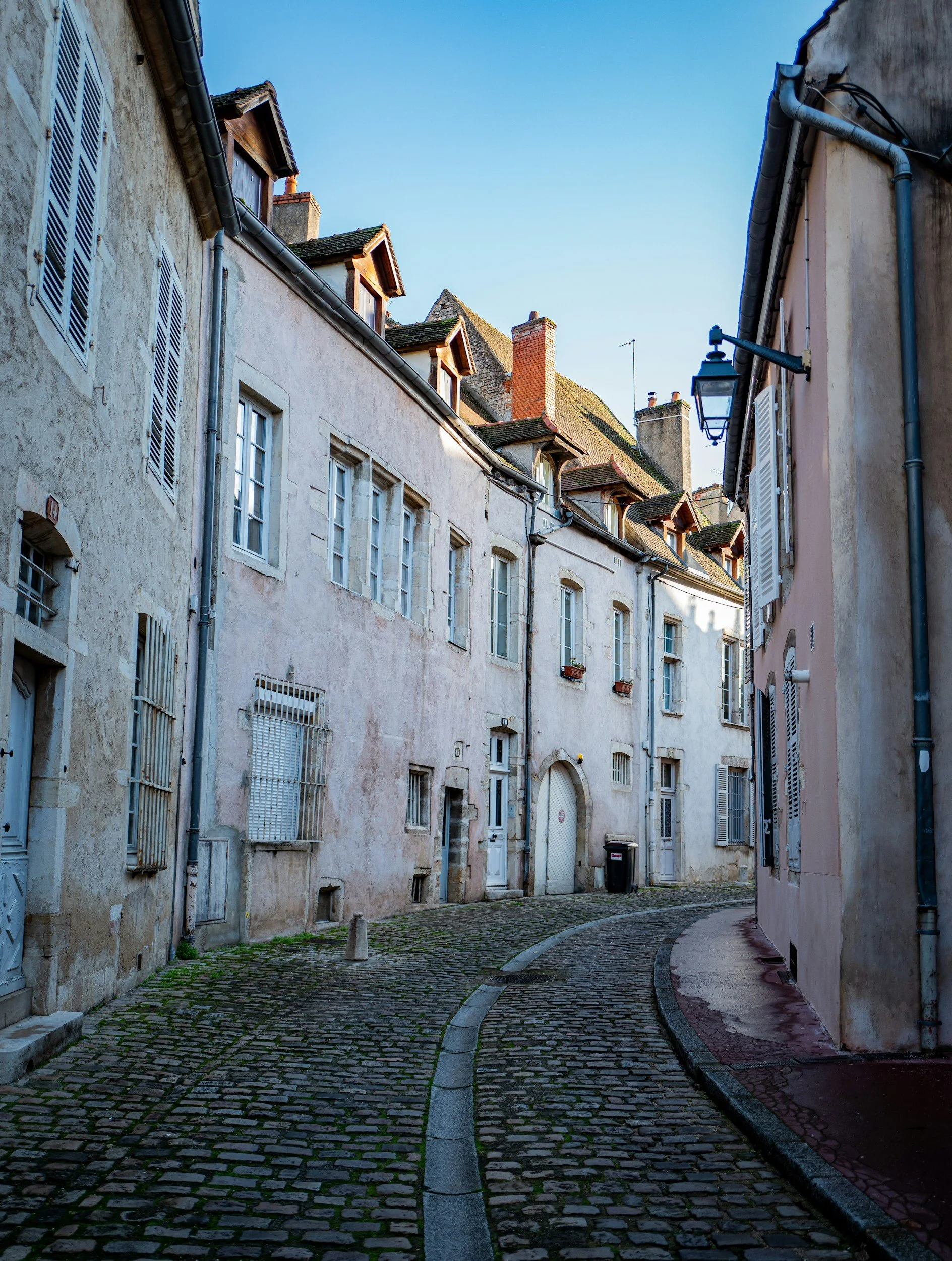 Ruelle pavée bordée de vieilles maisons avec fenêtres à volets et toits en pente, sous un ciel bleu.