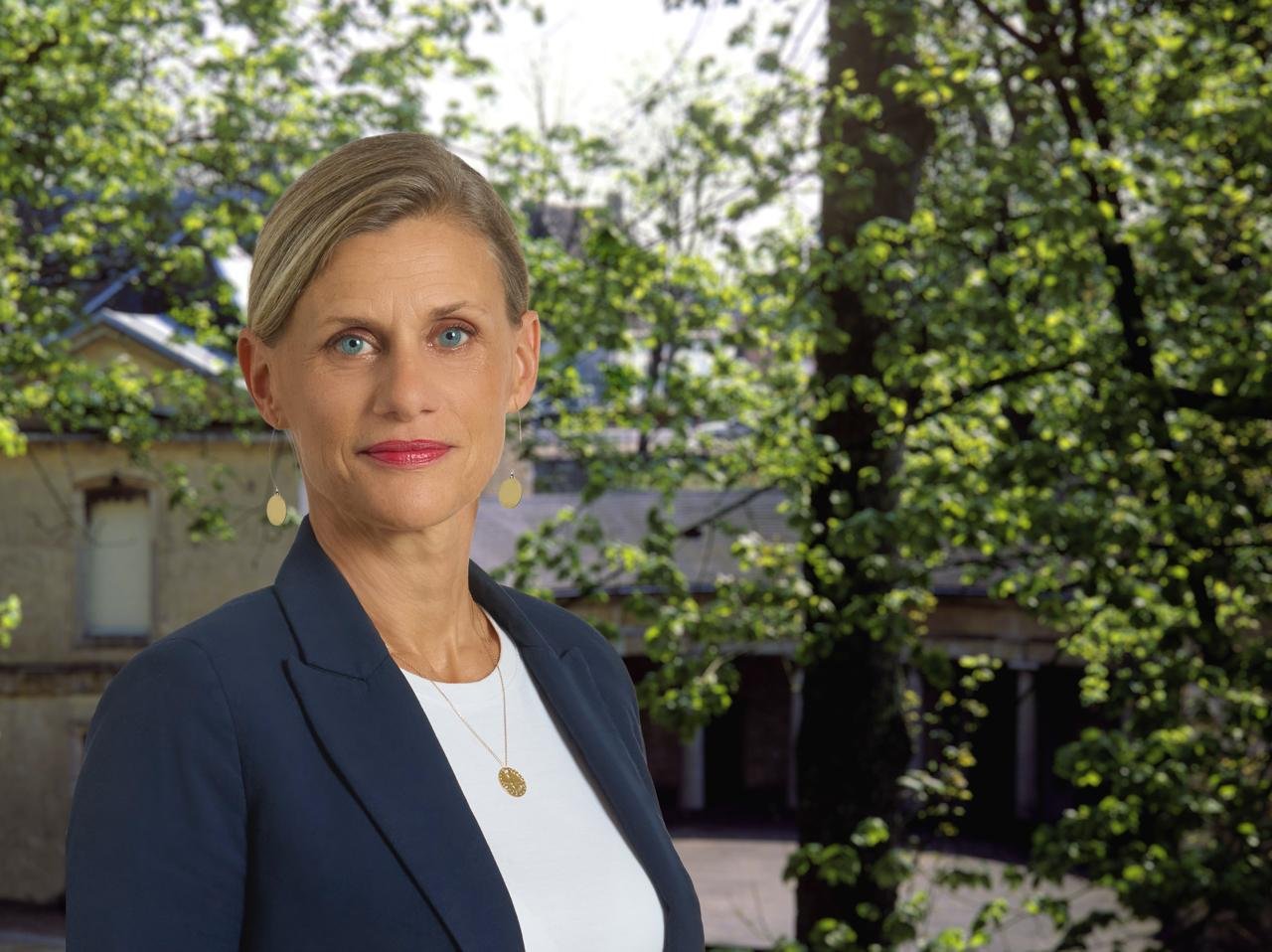 Femme aux cheveux courts et blonds, portant une veste bleue et un t-shirt blanc, se tenant devant un arrière-plan de arbres en plein air.