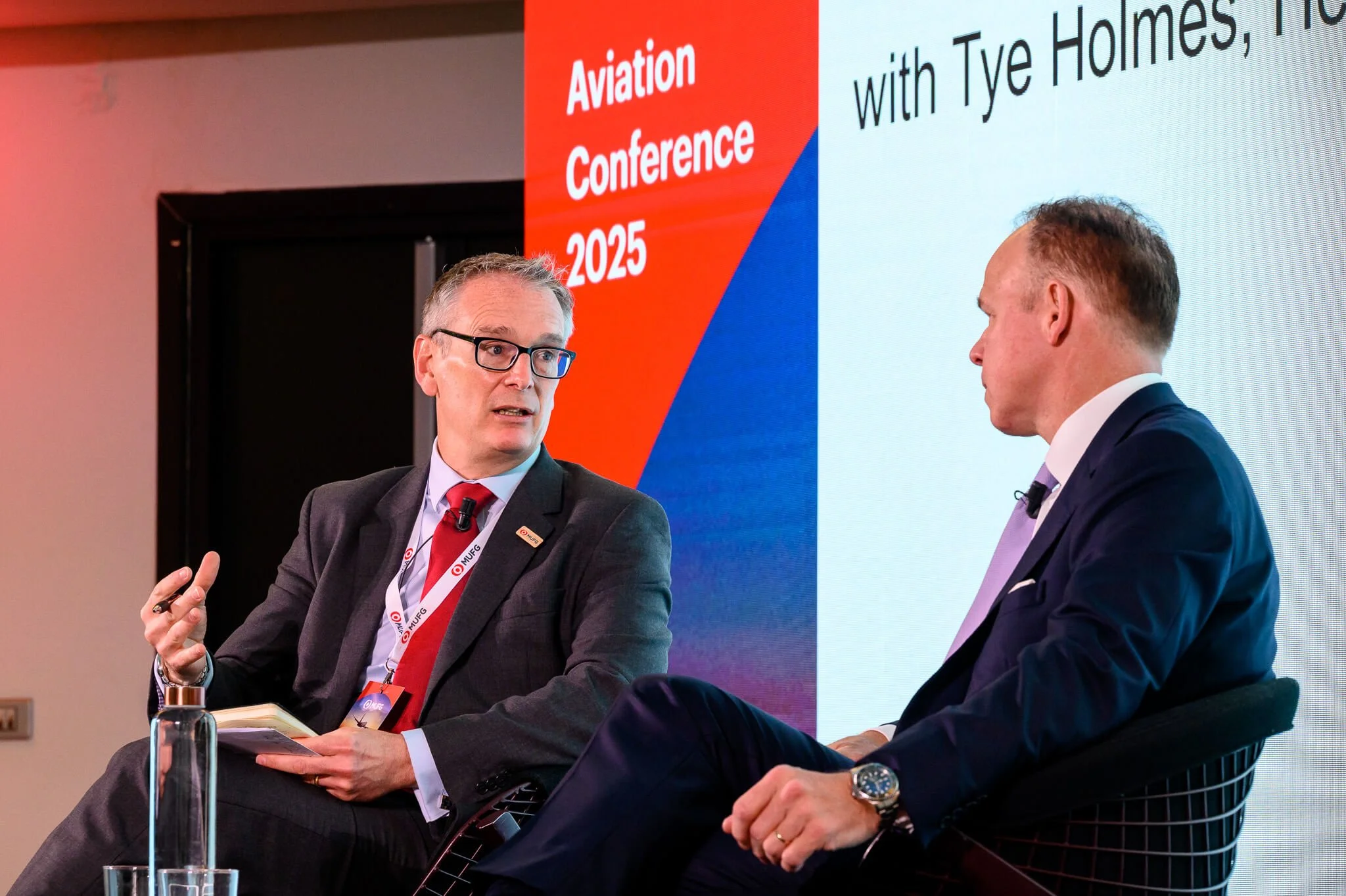 Two men in suits and ties having a discussion on stage at the Aviation Conference 2025, with a large screen behind them displaying the event's name and date.