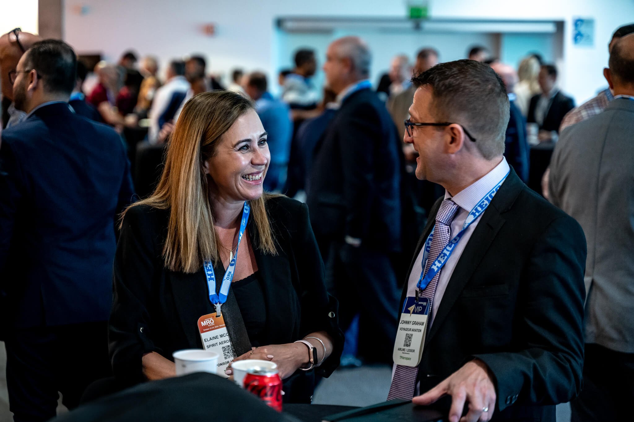 Two professionals, a woman and a man, are smiling and engaging in a conversation at a networking event or conference with many people in the background.