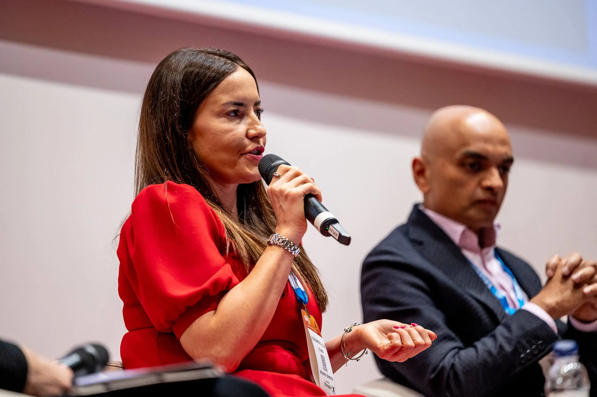 A woman in a red dress speaking into a microphone during a panel discussion, with a man in a dark suit sitting beside her.