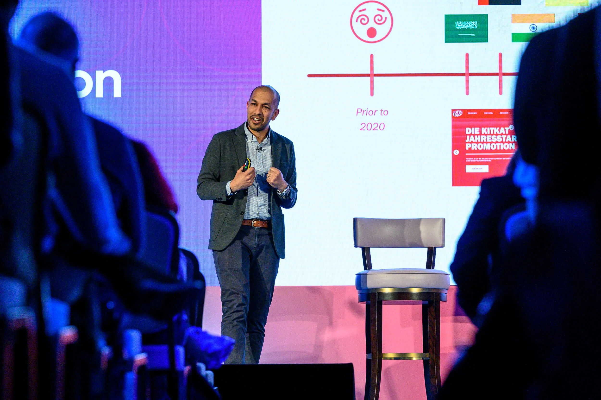 A man is giving a presentation on a stage with a large screen behind him, showing a timeline and text. The audience is seated and listening.