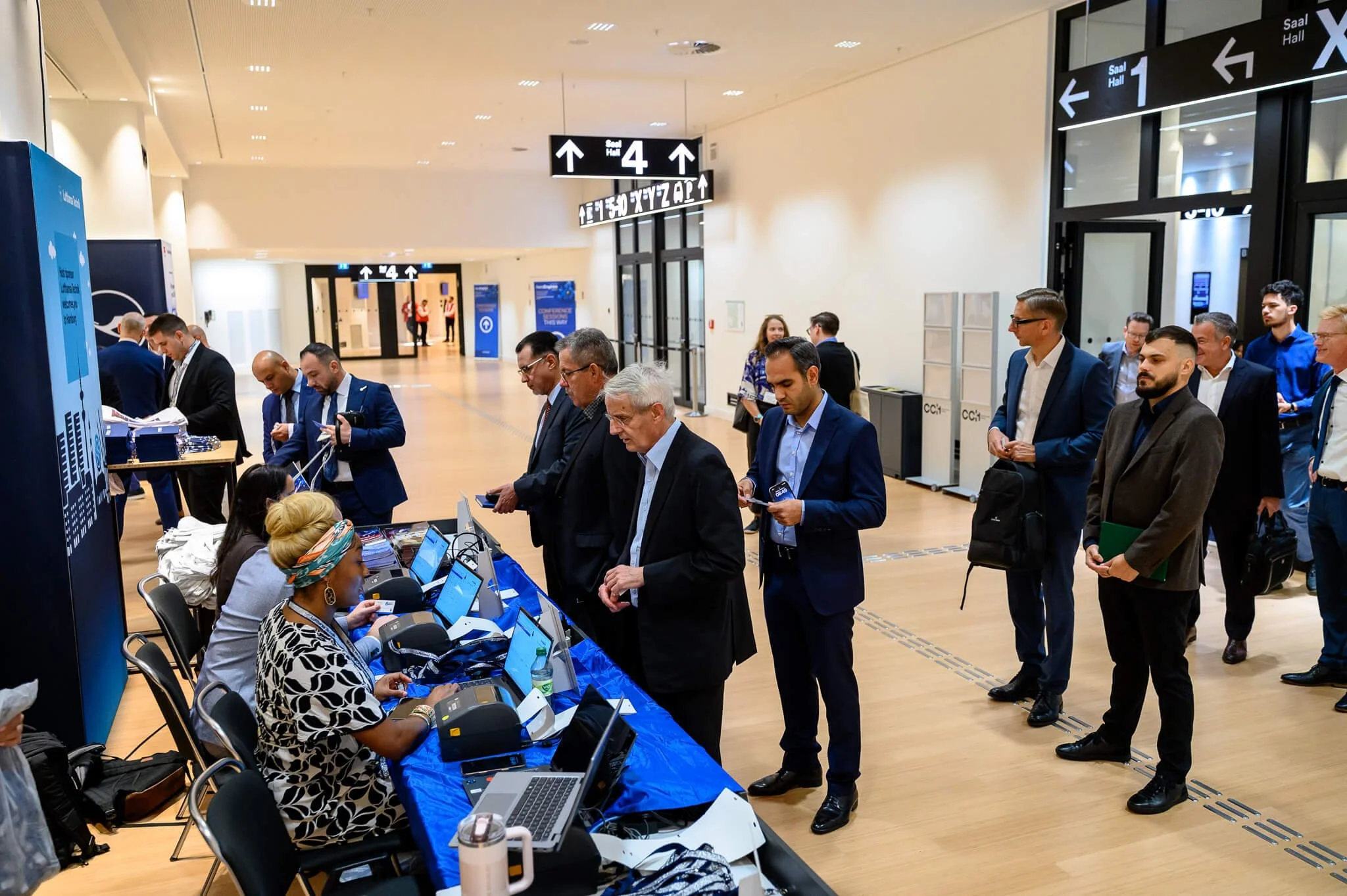 People in business attire registering at a conference check-in desk in a modern, spacious venue with directional signs overhead.