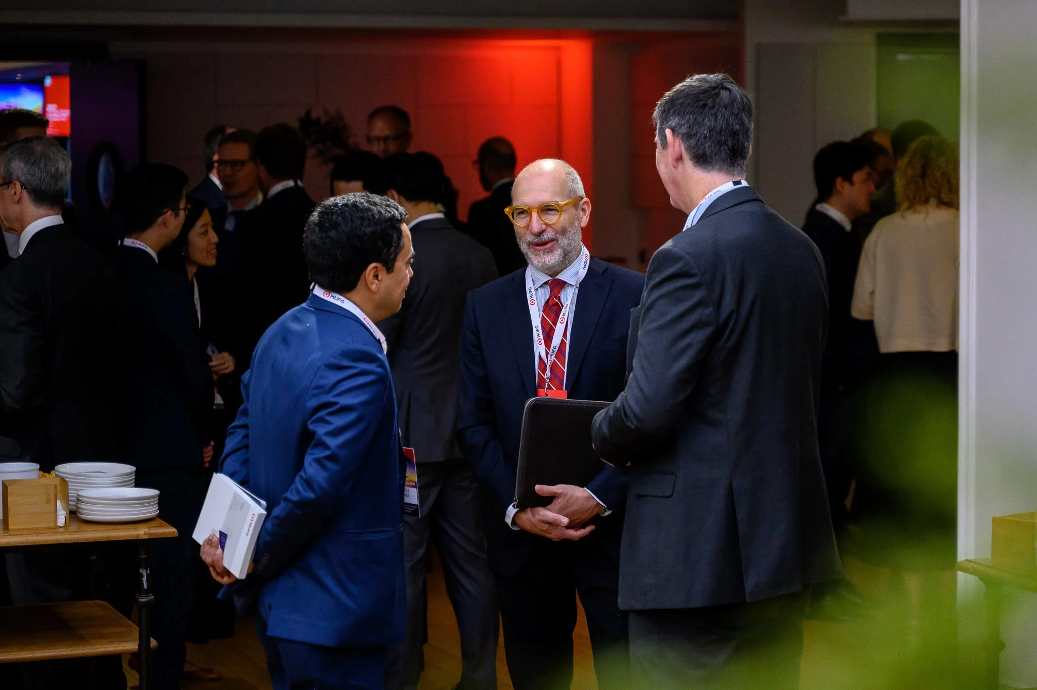 Three men in suits engaged in conversation at a professional networking event, with more attendees in the background.