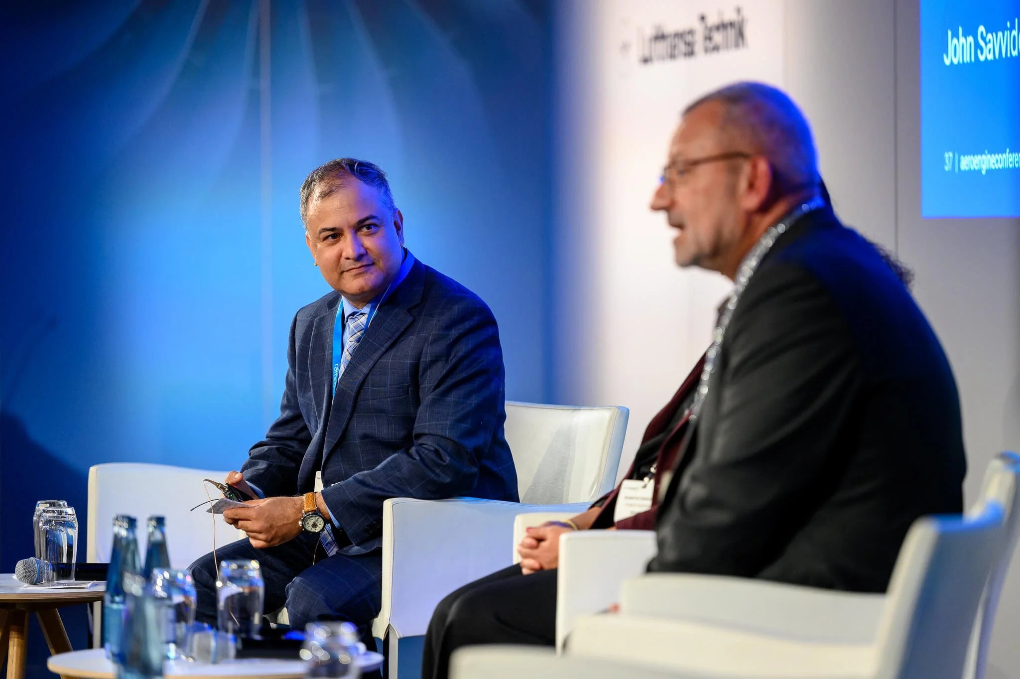 Two men in business attire sitting on a stage at a conference. One man is looking at the other, who is speaking. There are glasses of water and microphones on small tables in front of them.