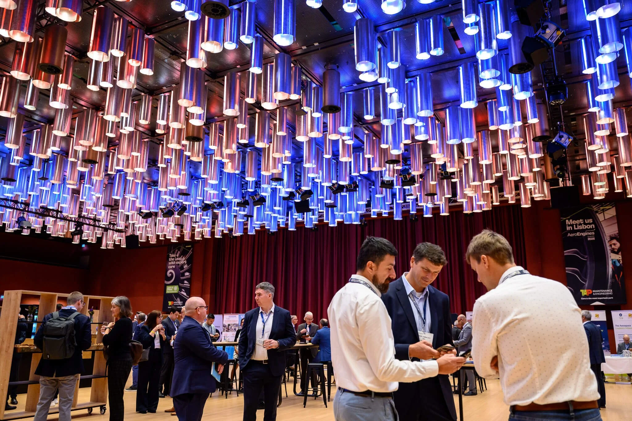 Conference or networking event with people in business attire, some using phones, in a room with a decorative ceiling featuring numerous hanging, cylindrical blue and metallic pipes and multiple spotlights.