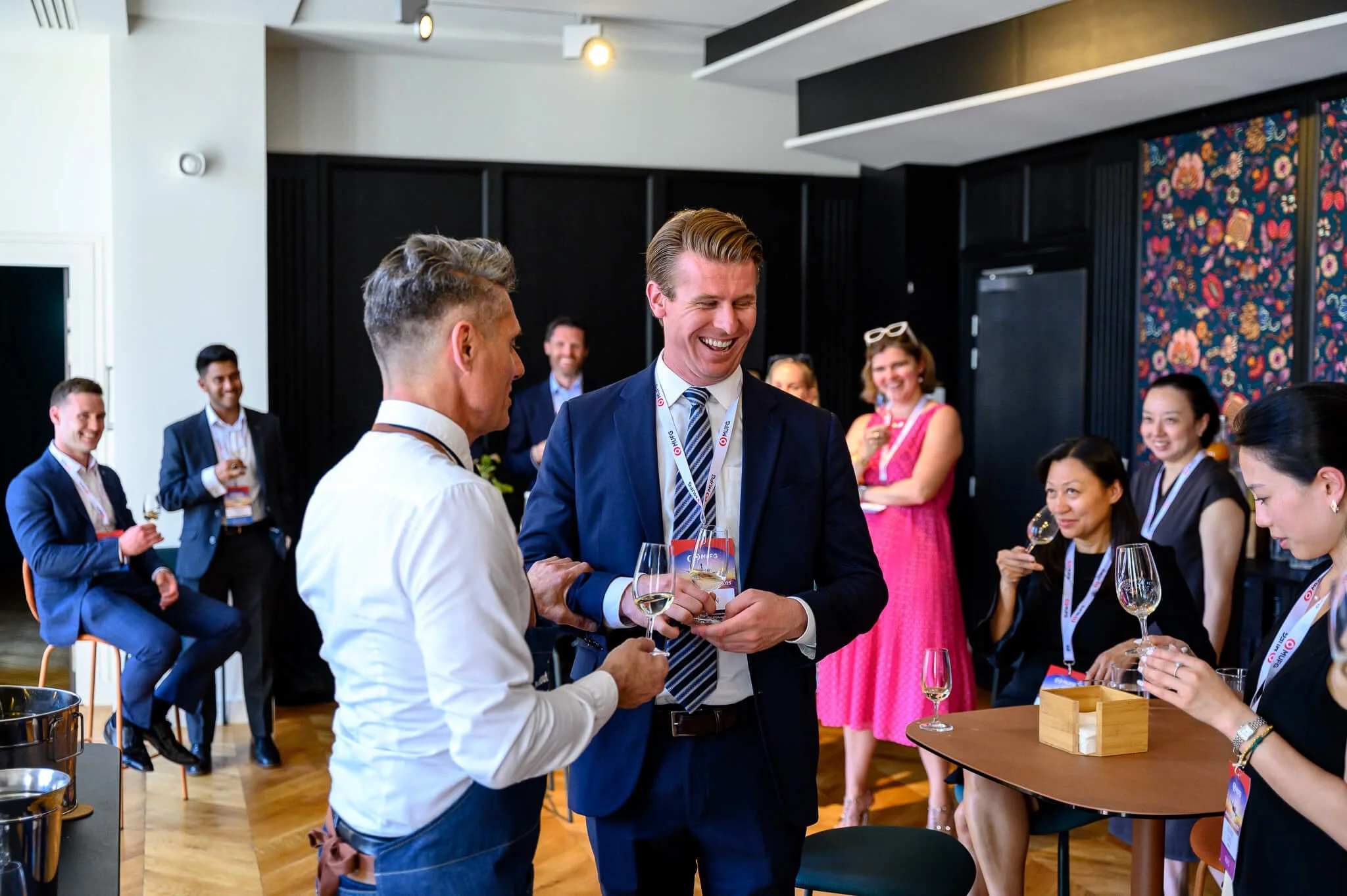 Business professionals at a networking event, holding wine glasses and engaging in conversation, in a modern, well-lit room.