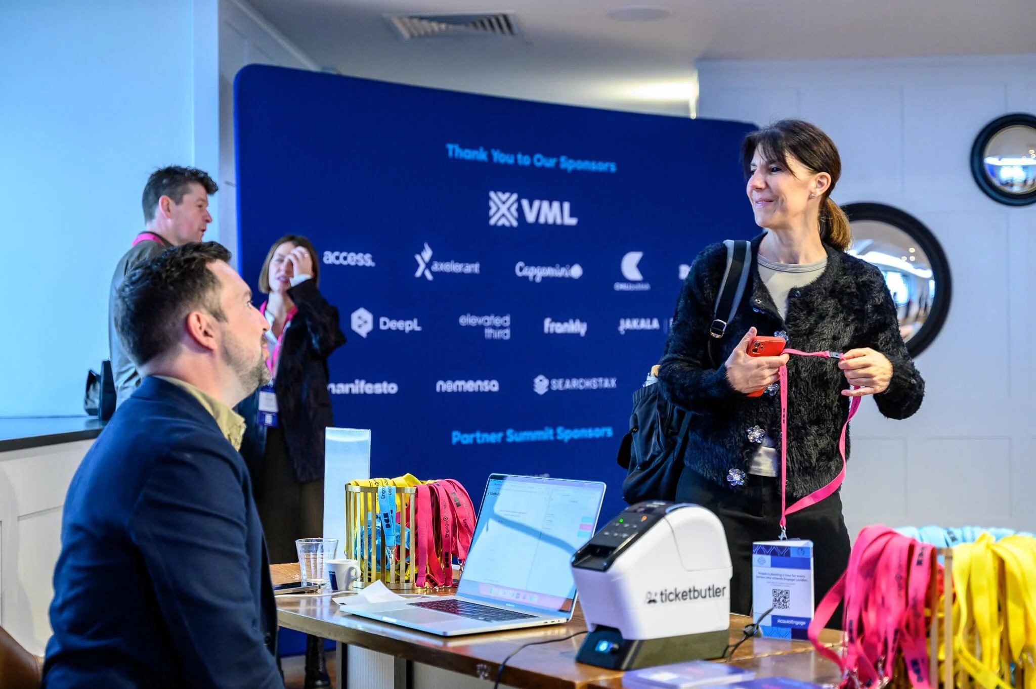 Conference registration area with a woman holding a phone and pink lanyard, standing at a table with a laptop, badge, and ribbons, engaging with two seated men. A large blue banner with sponsor logos is in the background.