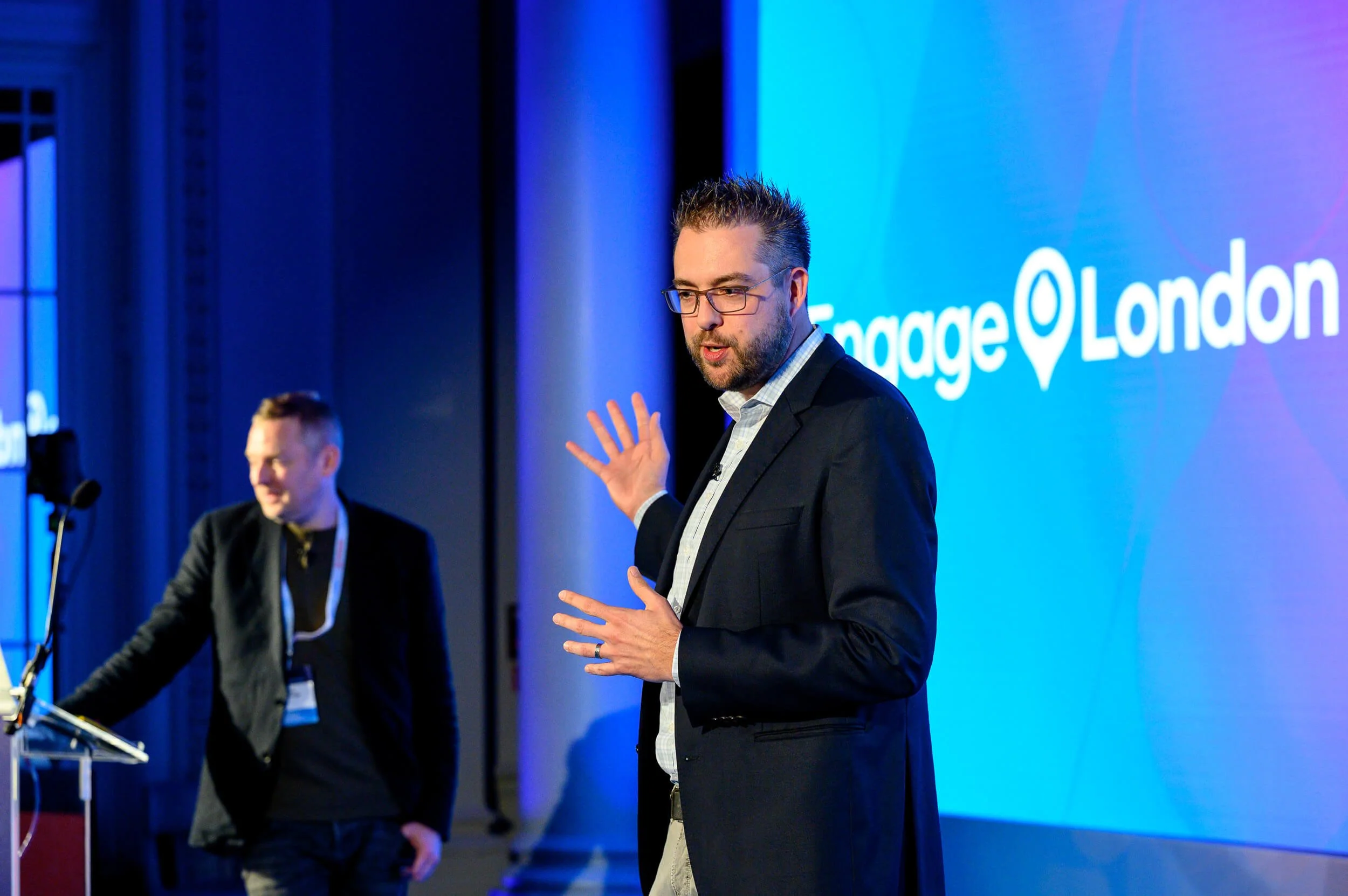 Two men speaking at a conference with a blue background displaying the words 'Engage London' and a location pin icon.