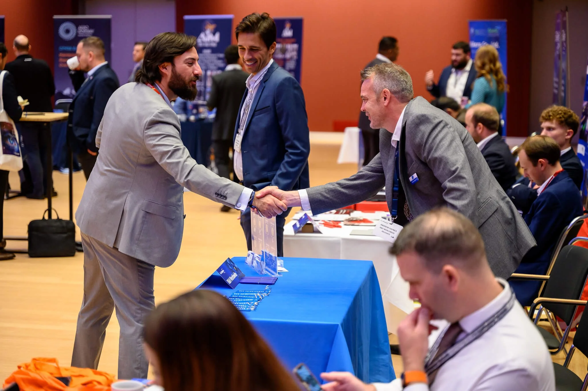 Two men in suits shake hands at a conference booth, smiling and engaging in conversation, while others sit and stand around booths and tables in a conference room.