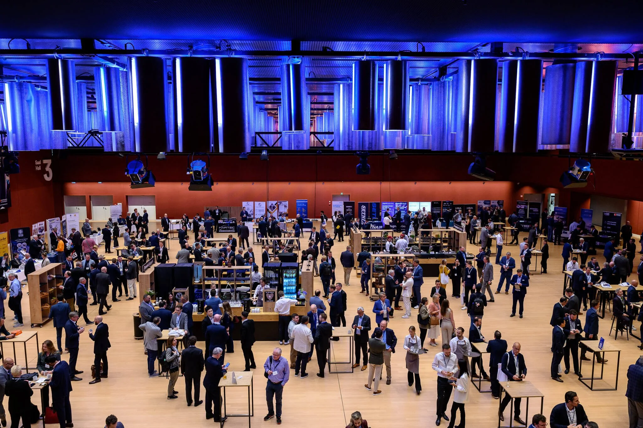 A large conference or trade show with many people walking around and interacting. Attendees are dressed in business attire, and there are exhibition booths and displays throughout the space. The room has a high ceiling with blue and purple lighting.