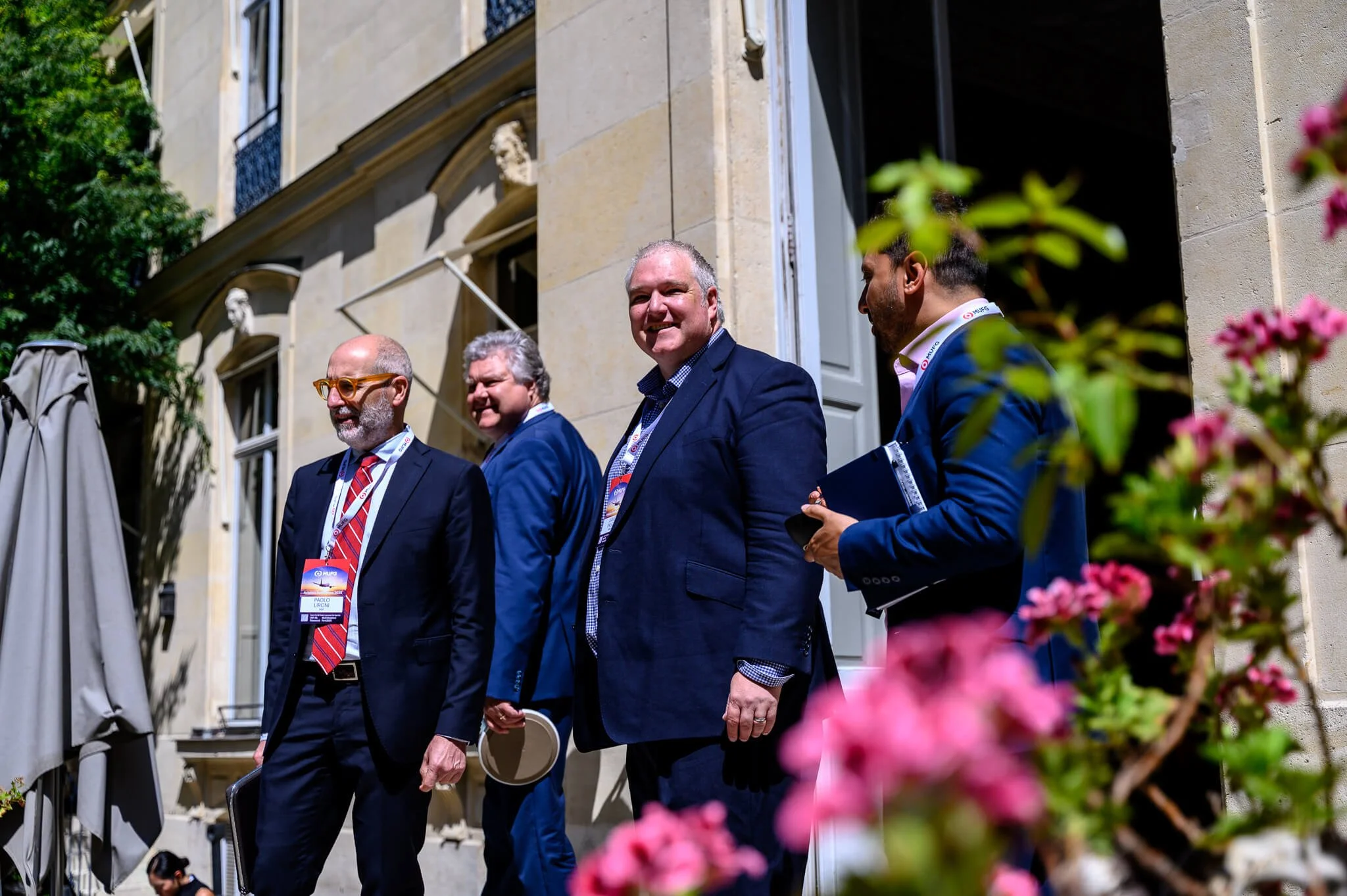 Four men in suits standing outdoors on a sunny day, some holding notebooks and wearing conference badges, near a historic building with flowers in the foreground.
