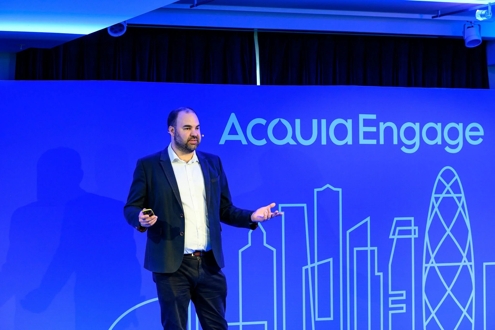 Man in dark suit giving a presentation at Acquia Engage conference, standing in front of blue backdrop with city skyline outline.