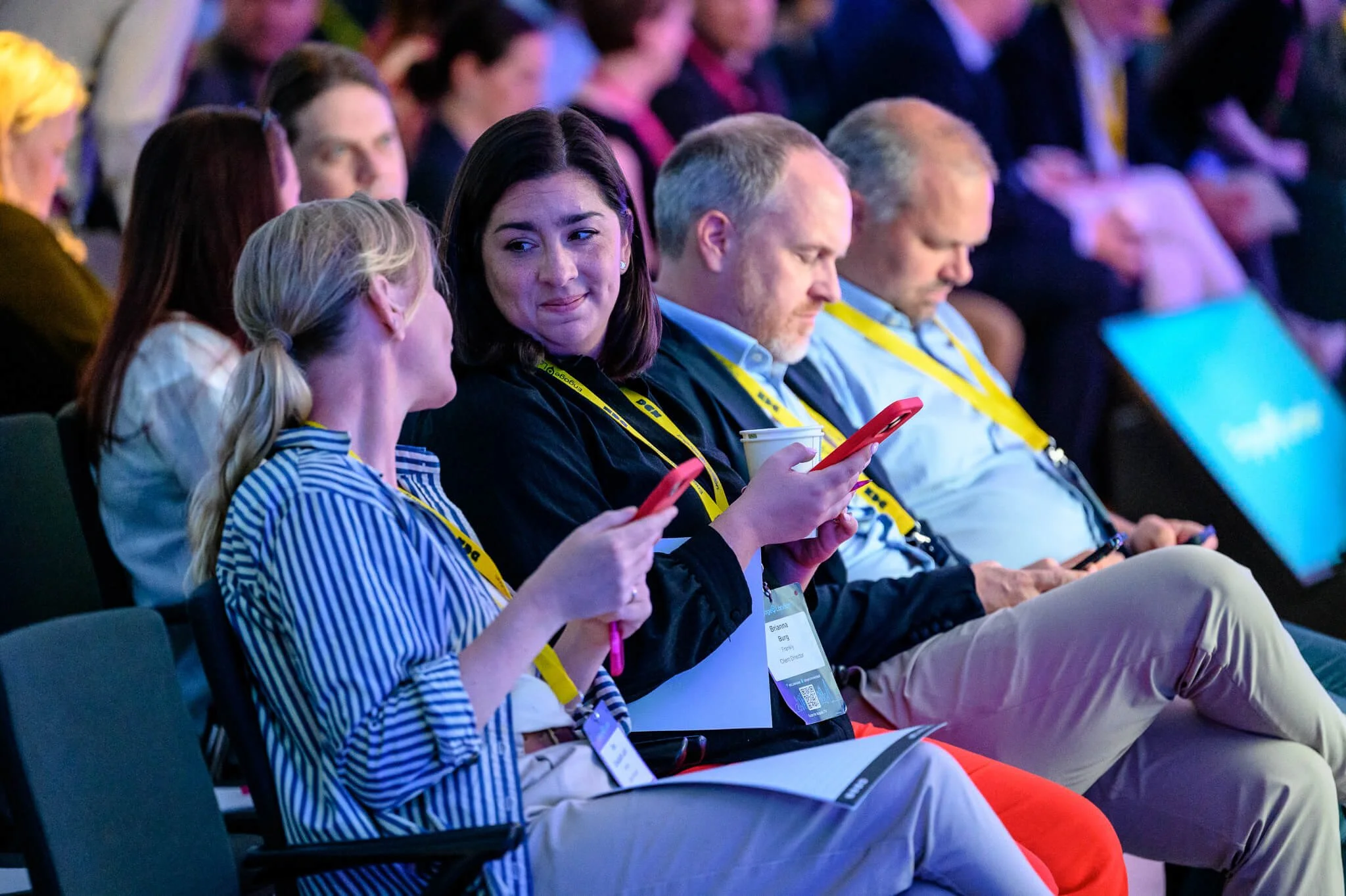 Group of people sitting in an audience at a conference or event, with some looking at their phones and others listening.