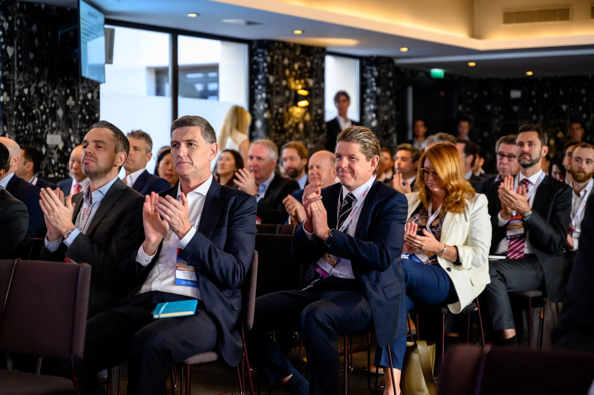 Business professionals attending a conference, seated and clapping, in a brightly lit modern venue with large windows.
