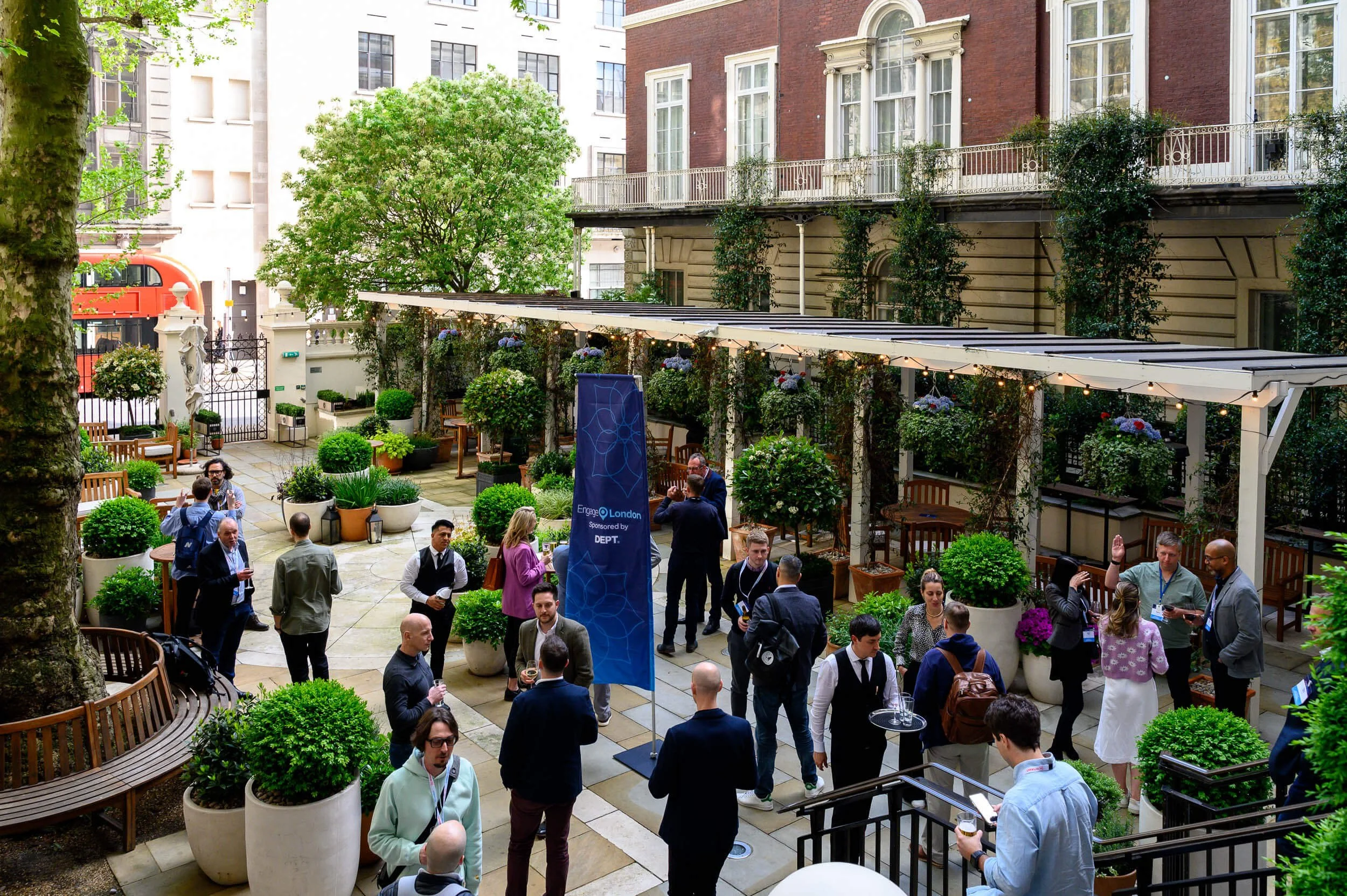 People gather in an outdoor garden area with potted plants and trees, under a canopy, next to a brick building. Some people are talking, holding drinks, or using their phones, at a social event.