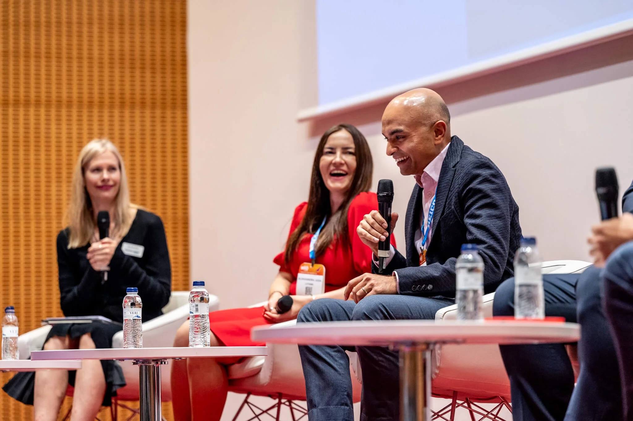 Three people on a panel discussion, with two women and one man, sitting on stage with water bottles and microphones, laughing and engaging in conversation.