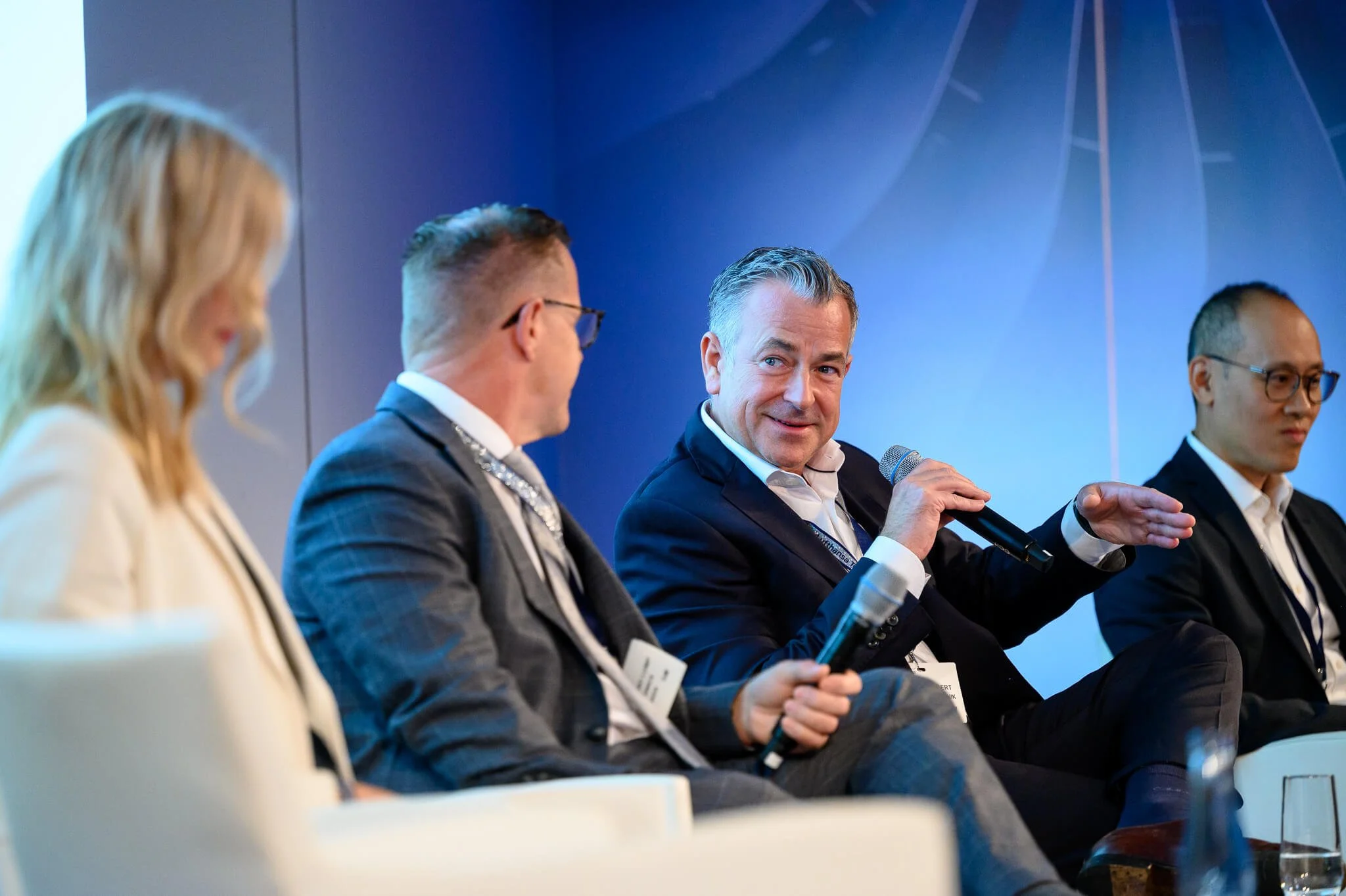 Group of four people, three men and one woman, sitting on stage participating in a panel discussion at a conference or event. The man in the middle is speaking into a microphone.