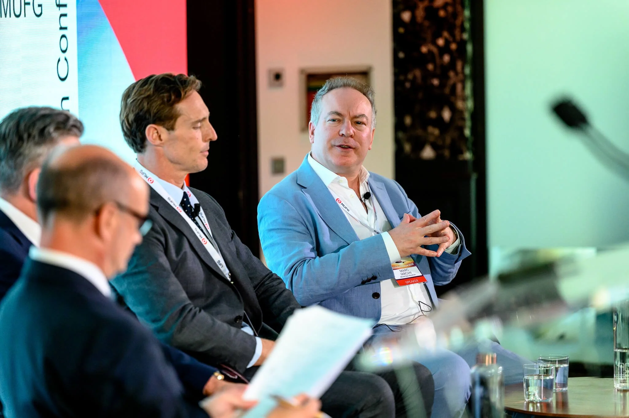 A panel of men seated during a conference, with one man in a light blue suit speaking while others listen, in an indoor setting with a backdrop displaying the logo 'MURE' and 'CONFERENCE'.