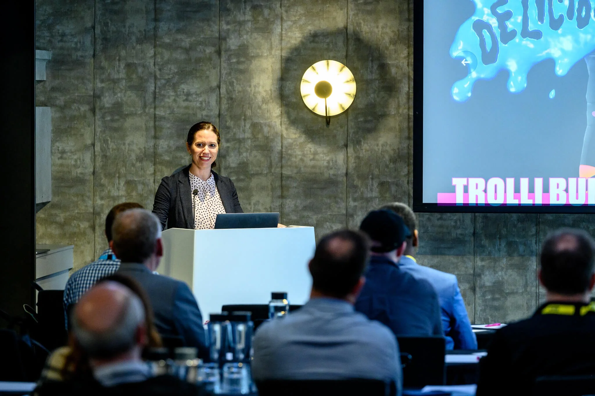 A woman presenting at a conference with an audience, a large screen displaying a graphic with the word 'DELICIOUS' and 'TROLLI BURST' visible in the corner.