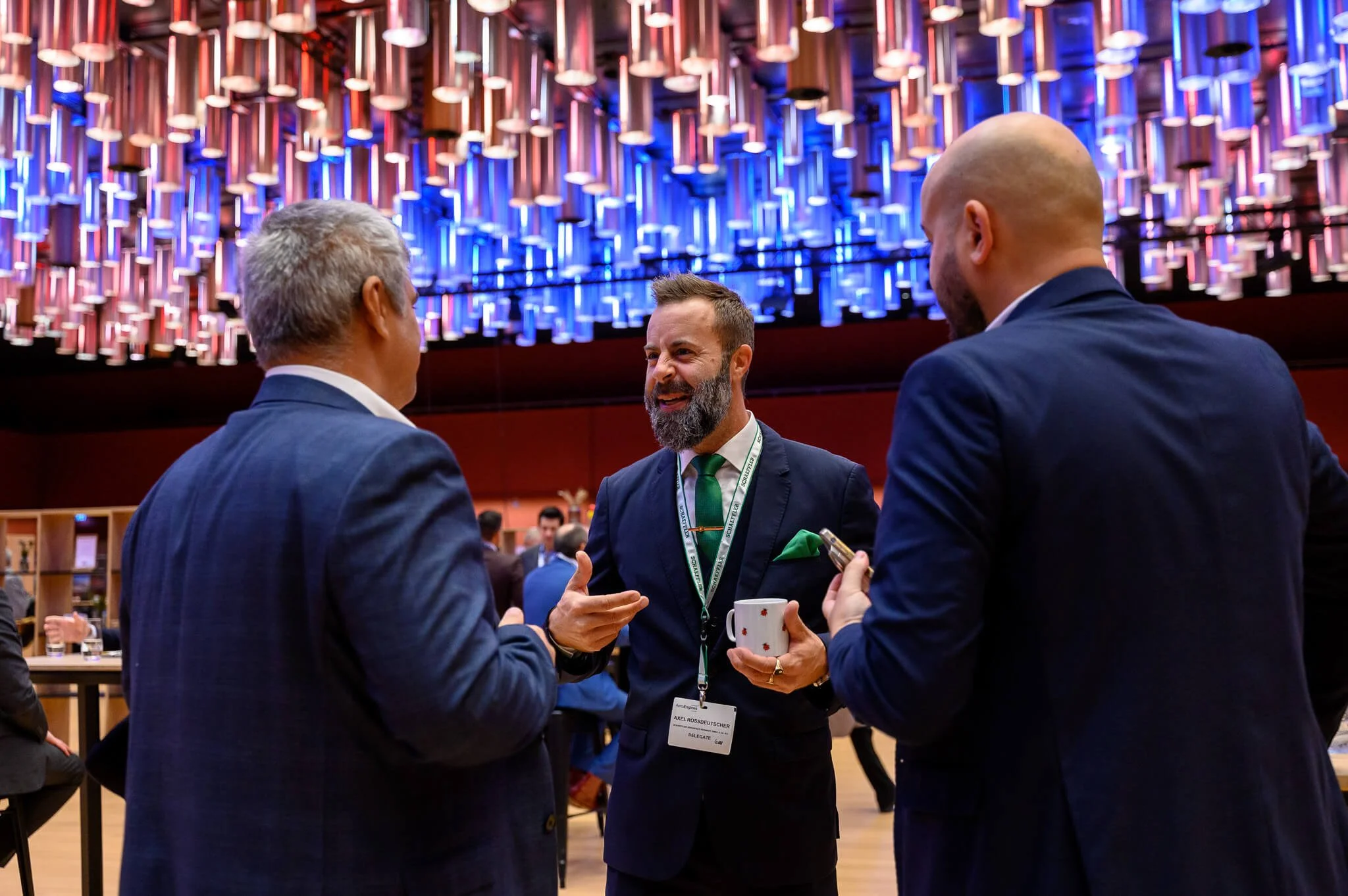 Three men in suits having a conversation at a professional event, with a vibrant, modern, colorful chandelier overhead.