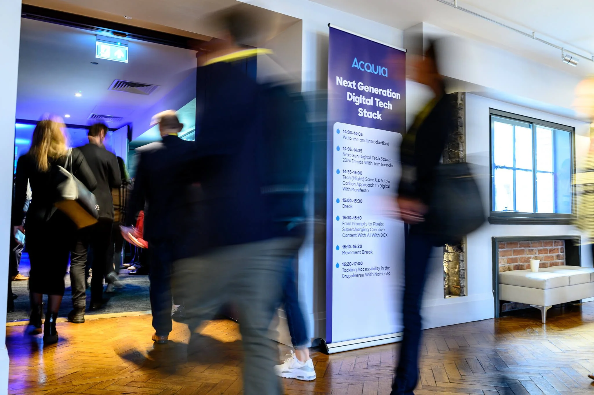 People walking through a conference event with a large schedule sign on the right, displaying various talks and sessions about digital technology, in a modern, well-lit room.