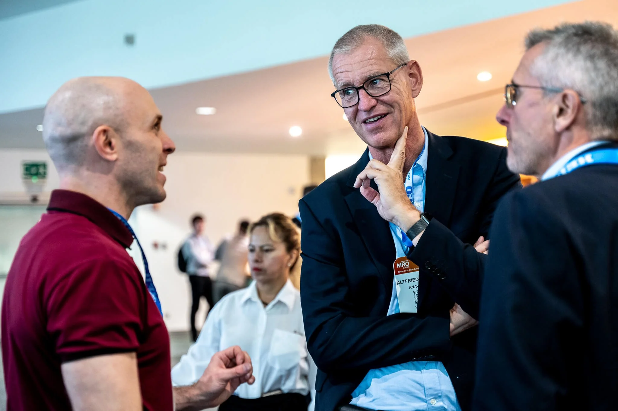 Three men and one woman engaged in conversation at a conference. The men are wearing suits and the woman is wearing a white blouse. The background shows a blurred conference hall with other attendees.