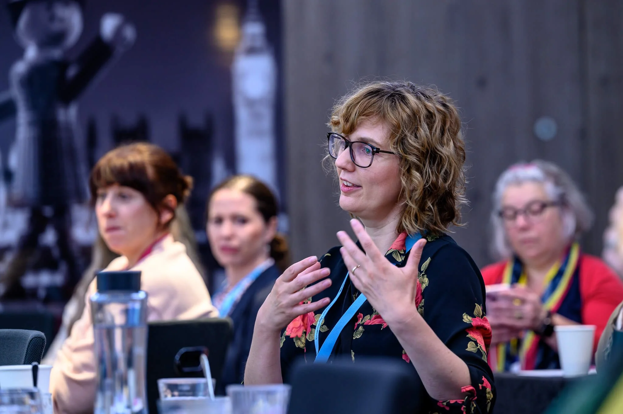 Woman with glasses speaking and gesturing with hands at a conference or seminar, seated at a table with other women, some of whom are looking at her and using their phones.
