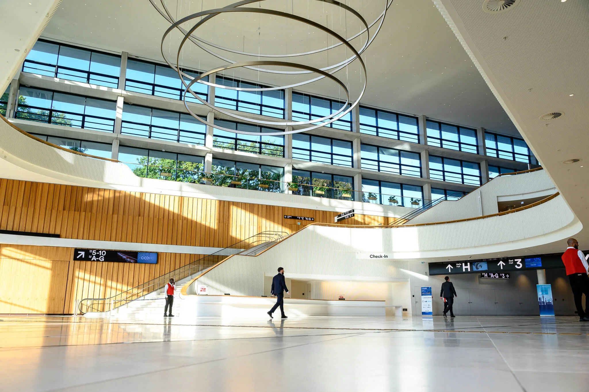 Interior view of a modern airport terminal with large glass windows, wooden accents, curved white railings, a spiral hanging light fixture, and people walking around.