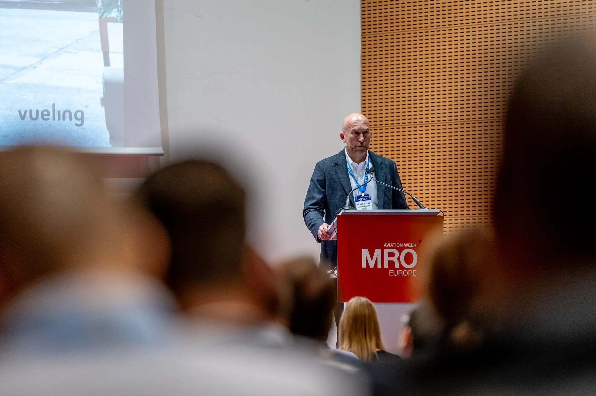A man speaking at a conference podium with a red sign reading 'AVIATION WEEK MRO EUROPE'. Audience members are visible in the foreground.