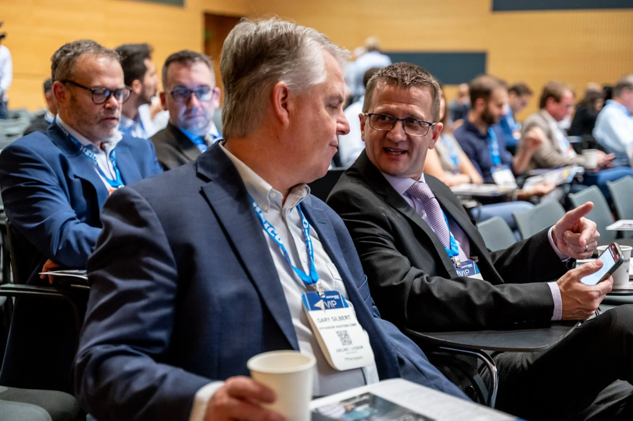 Two men in suits and glasses seated at a conference, engaging in conversation. One man is holding a smartphone and the other has a coffee cup. Both are wearing VIP conference badges.
