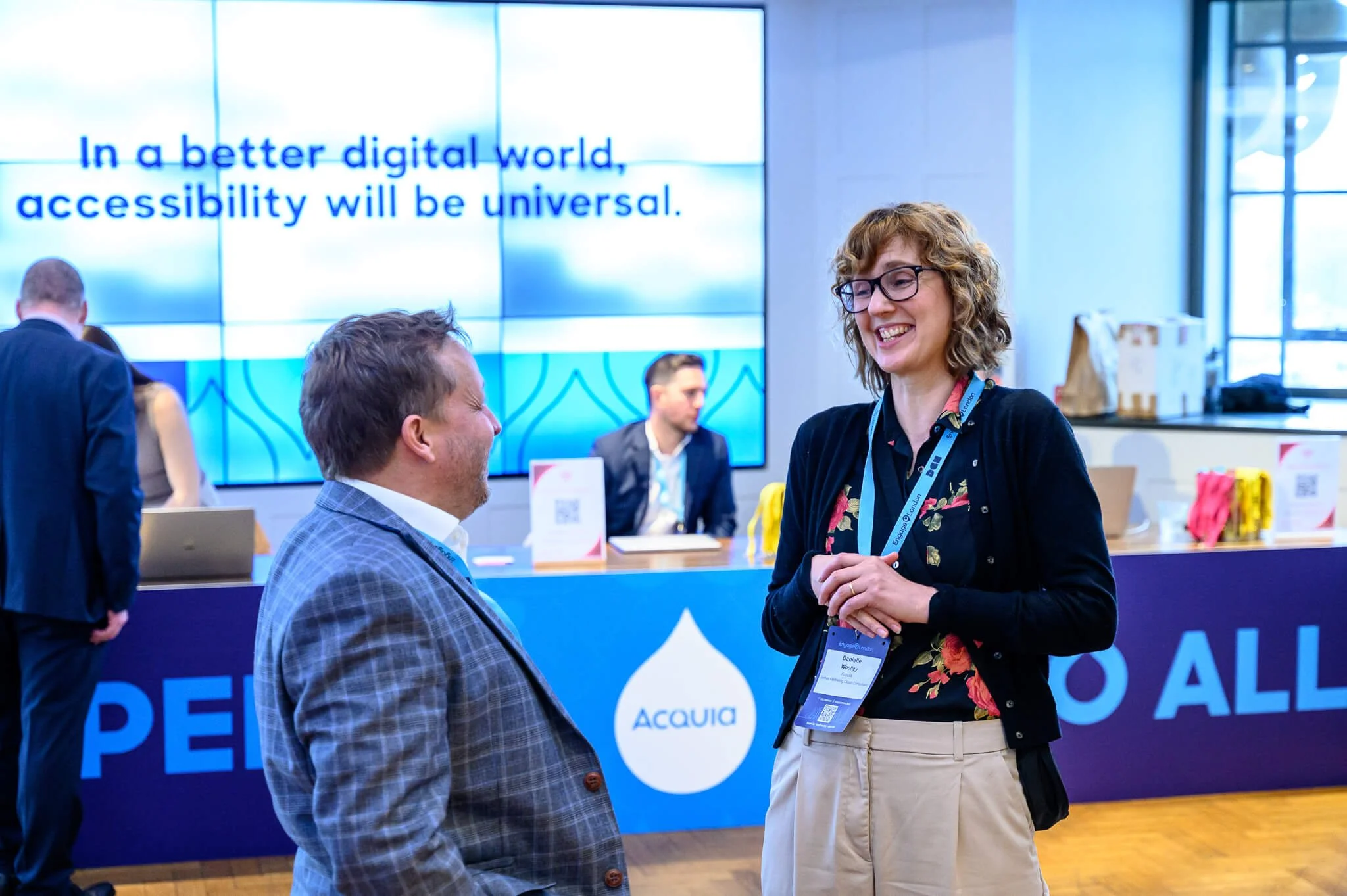 A man and woman are speaking and smiling at a conference behind a booth with signs and a large screen displaying the words "In a better digital world, accessibility will be universal." The woman has curly hair, glasses, a black cardigan, a floral blo