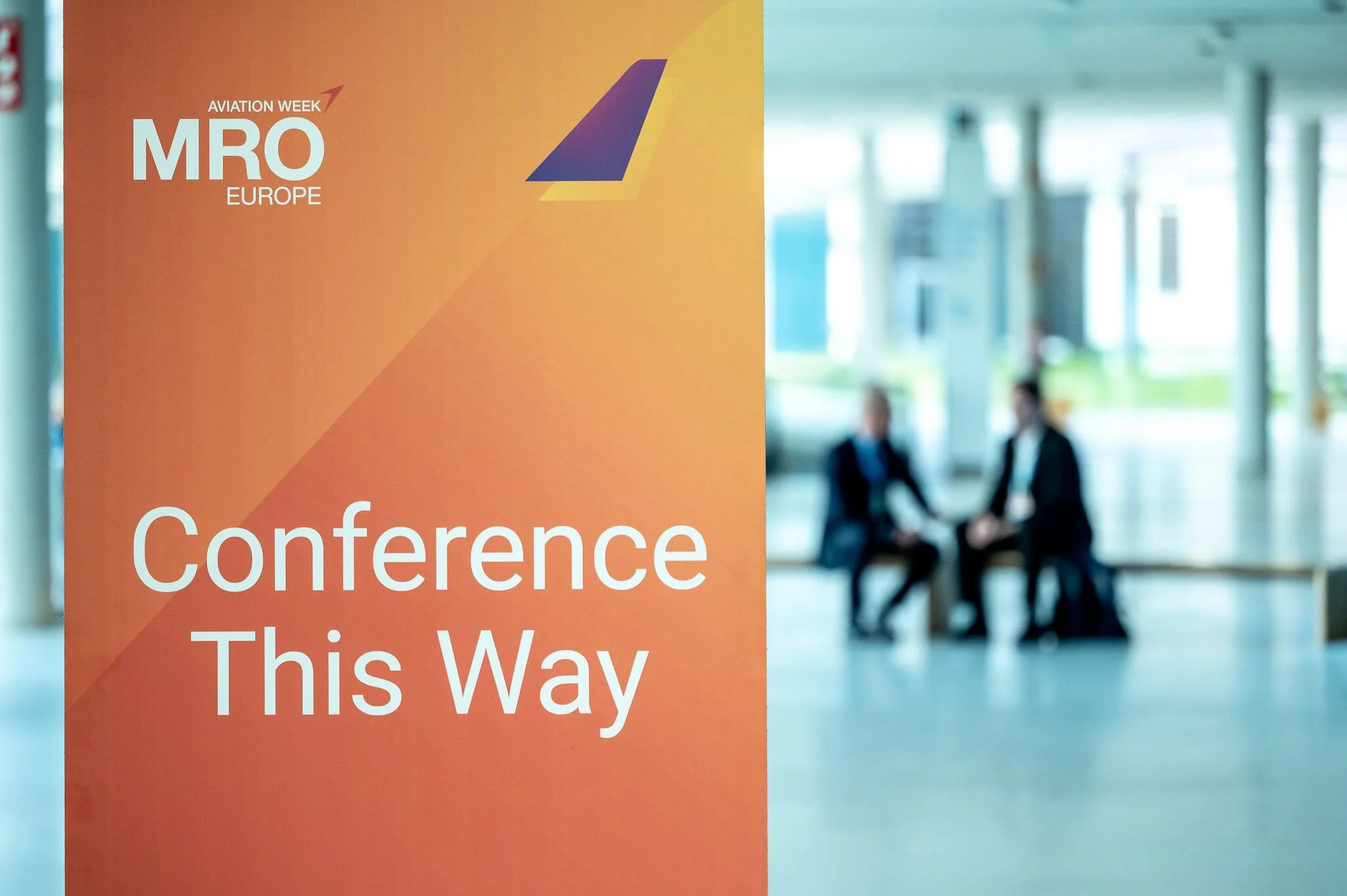 A large orange conference sign with text indicating 'Conference This Way' at an aviation event in Europe, with two blurred people sitting and talking in the background inside a modern building.