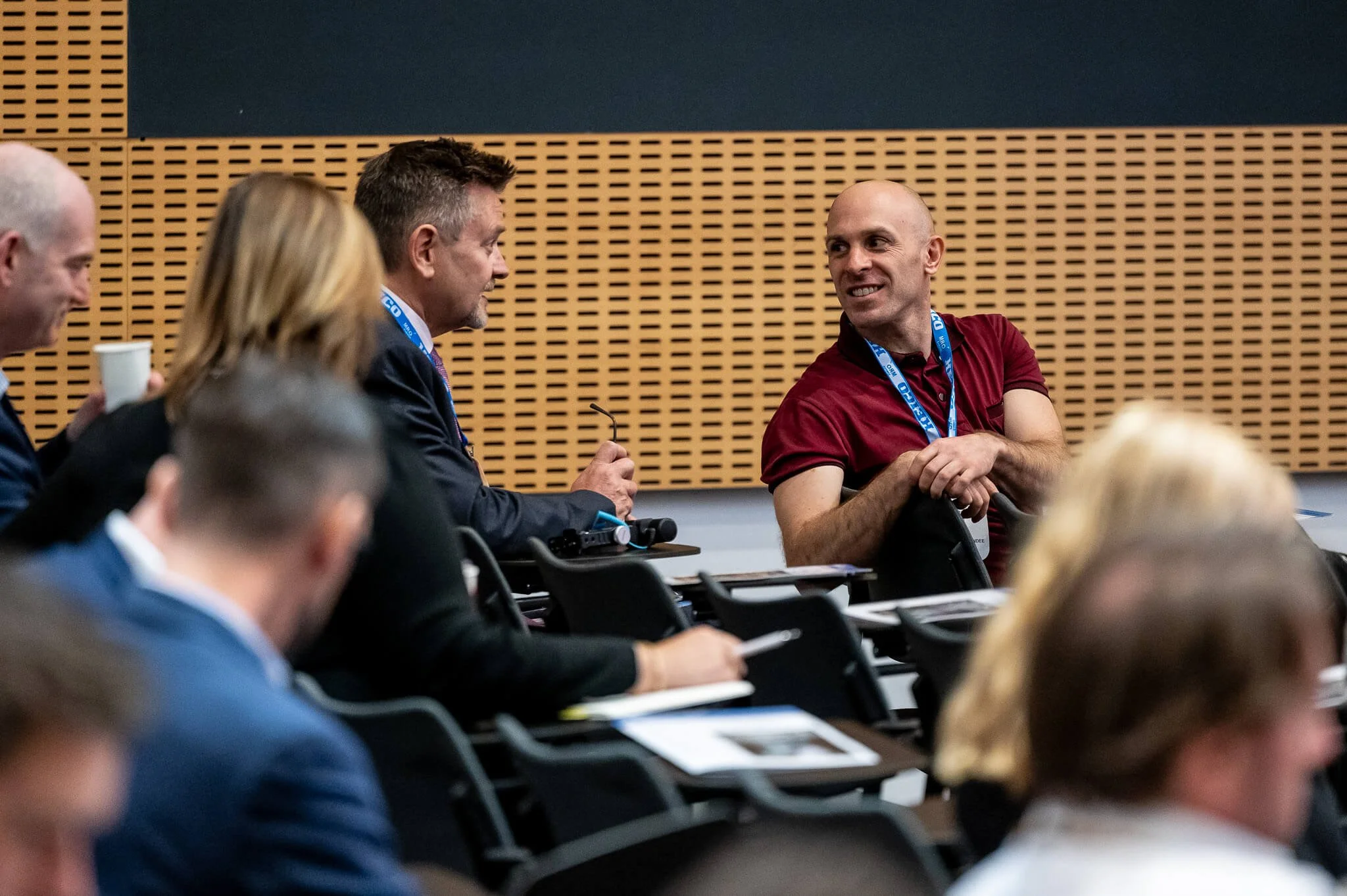 People attending a conference or meeting, some smiling and engaging in conversation, sitting at tables with papers and drinks.